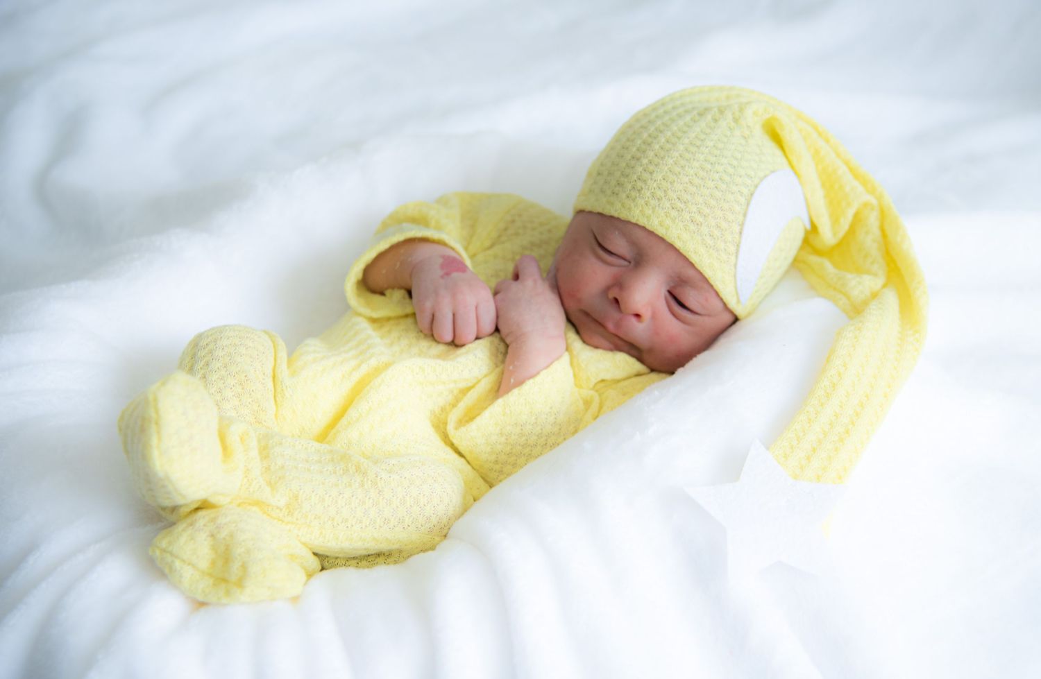 Sleeping newborn baby in yellow knit outfit and hat lying on white blanket.