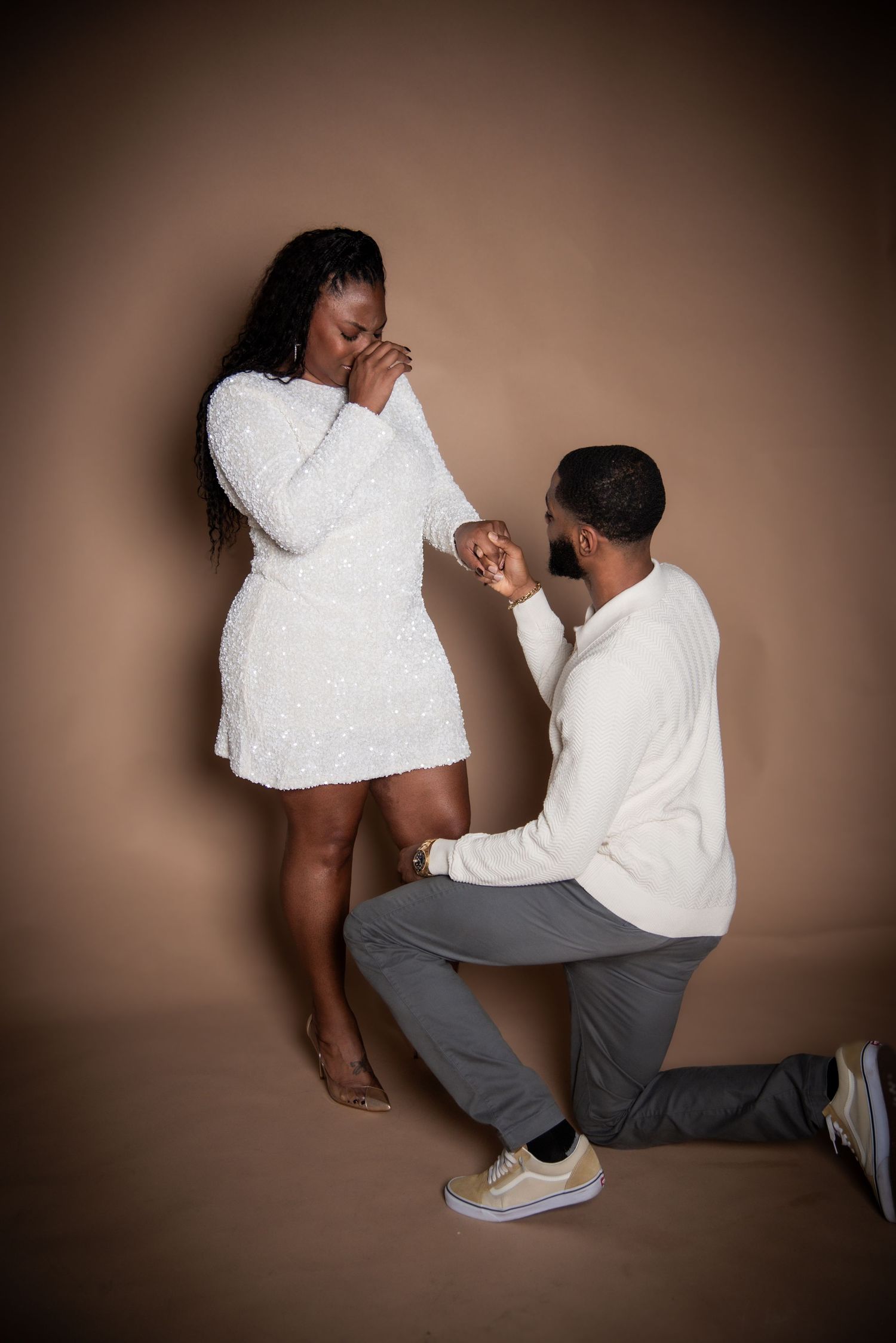 Man on one knee proposing to emotional woman in white sequin dress against warm brown studio background.