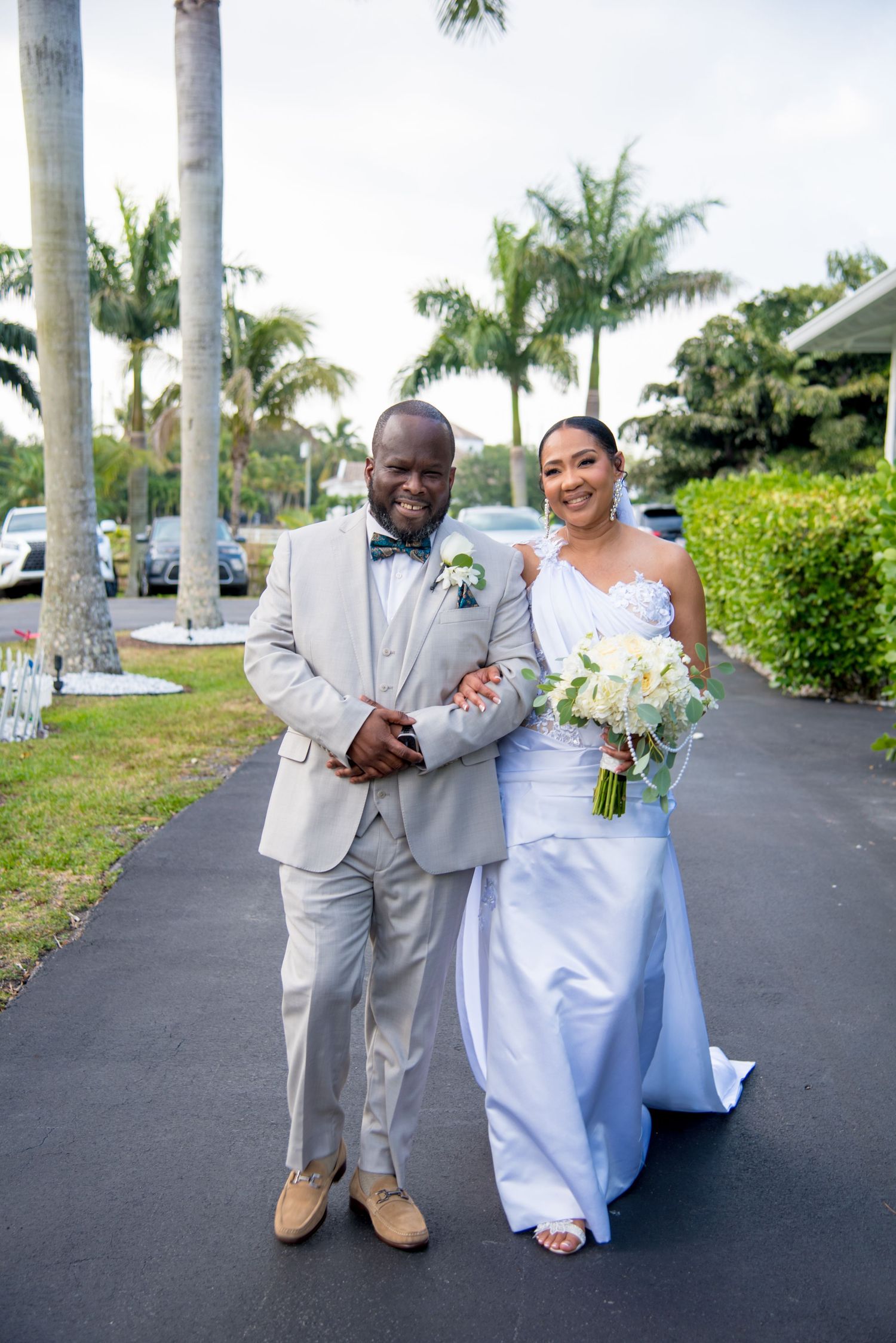 Bride in light blue gown and groom in gray suit pose together on a palm-lined path at a Florida wedding.