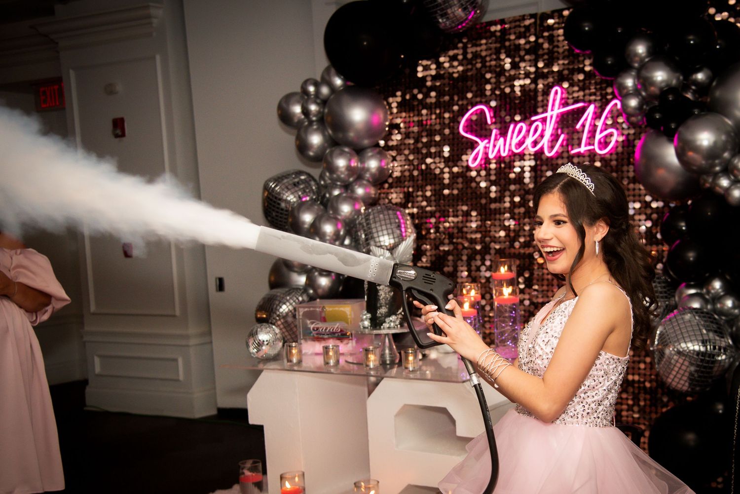 Teen girl in pink gown uses CO2 cannon at Sweet 16 party with balloon arch and neon sign backdrop.