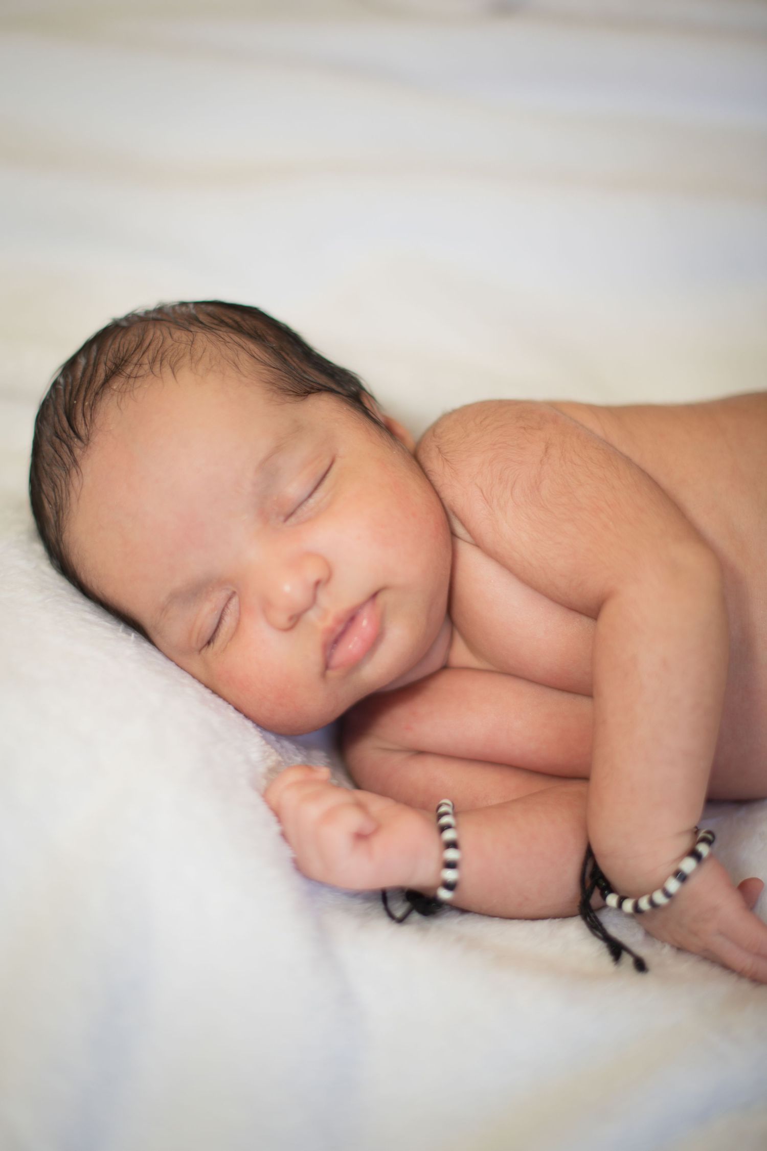 Peaceful newborn baby sleeping on white blanket wearing a black and white beaded bracelet on wrist.