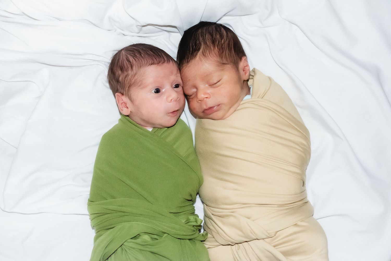 Two newborn twins swaddled in green and cream blankets sleeping peacefully side by side on white bedding.