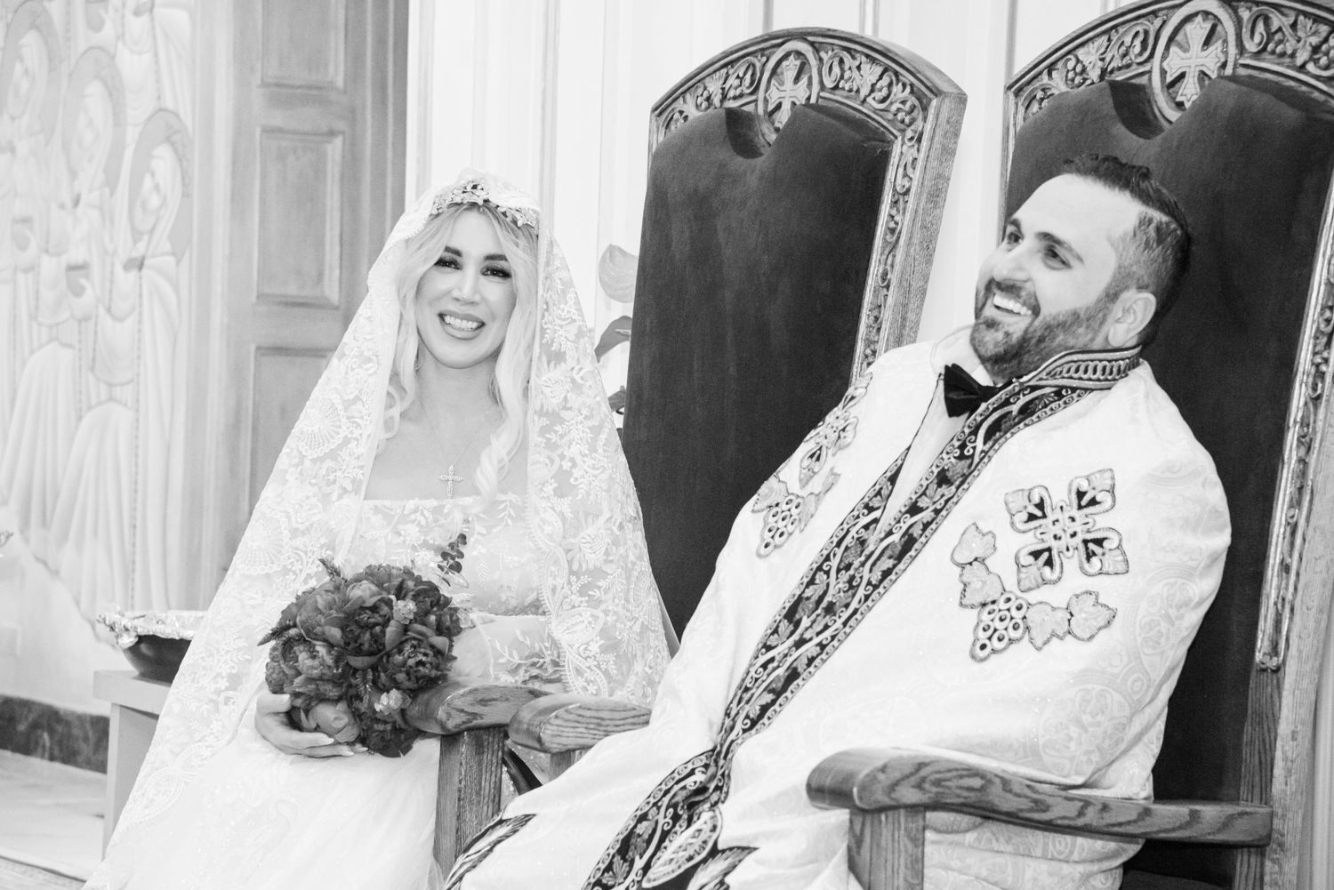 Black and white photo of a smiling bride in lace veil holding bouquet beside groom in ornate vestments at church wedding.