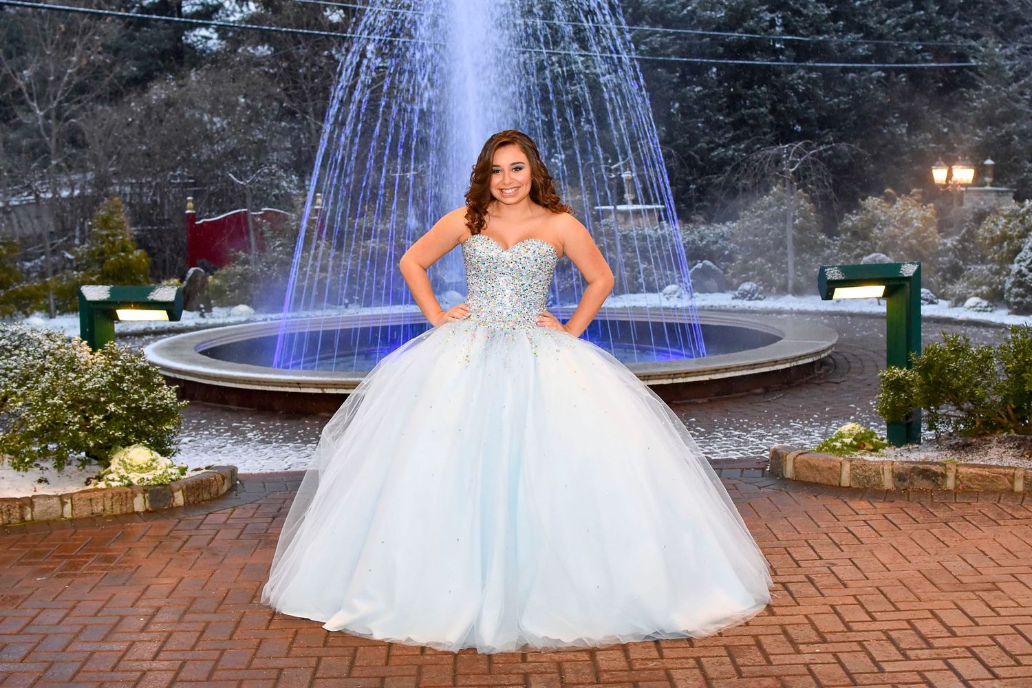 Young woman in light blue ballgown with jeweled bodice posing in front of illuminated fountain at dusk.