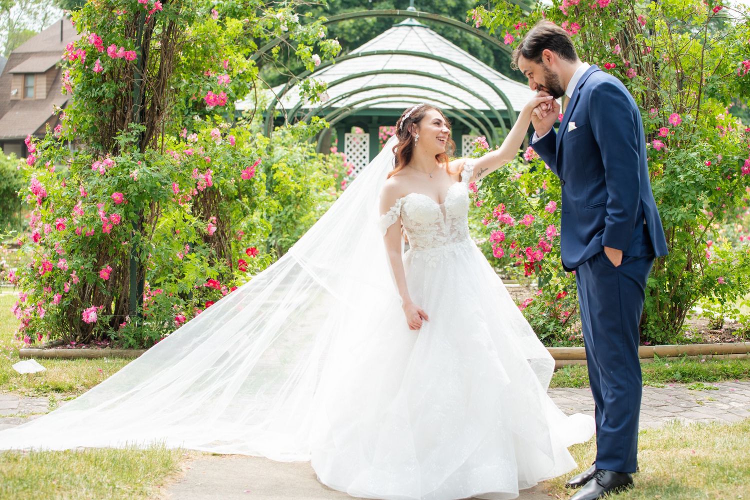 Groom in navy suit kisses bride's hand in white ballgown near floral garden gazebo on their wedding day.