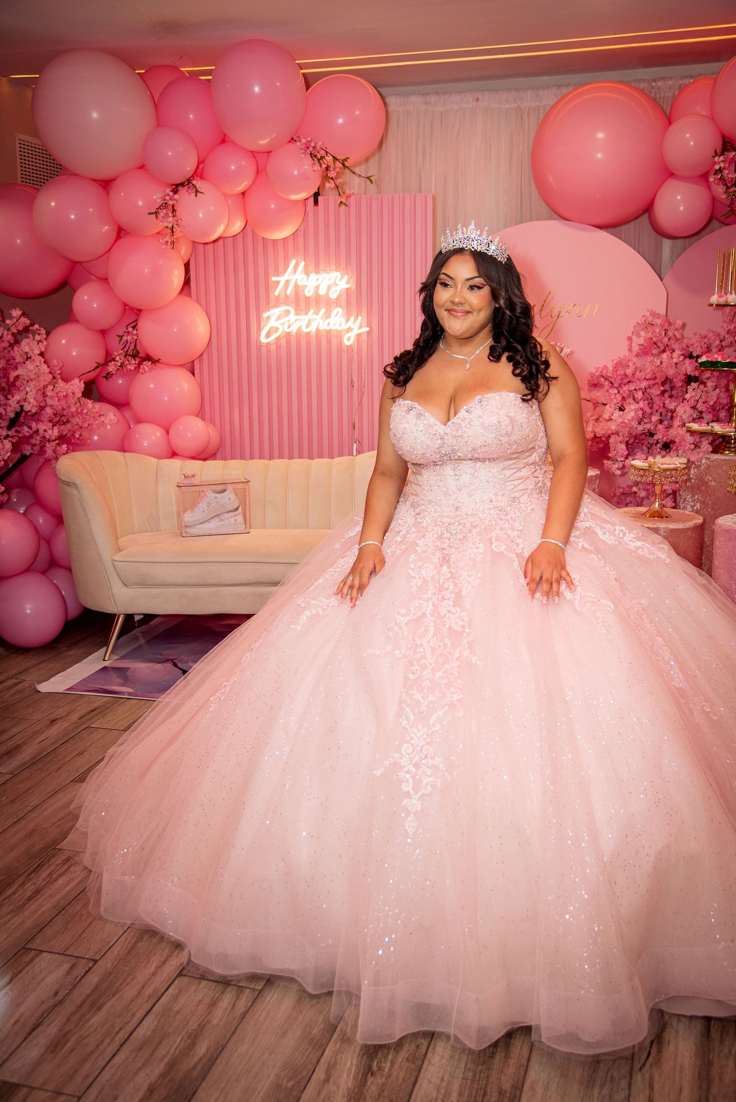 Young woman in pink ball gown and tiara poses at a pink-themed quinceañera party with balloon decor.