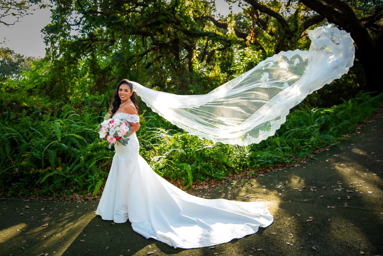 A flowing white wedding dress with a long veil catches sunlight in a lush garden setting by FBM Studios in Ft Lauderdale Fl.