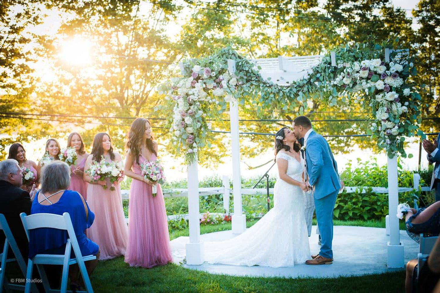 Couple shares first kiss at outdoor wedding ceremony under floral arch, with bridesmaids in pink dresses at golden hour by FBM Studios in FL