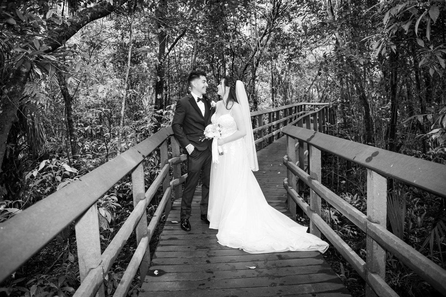 Black and white photo of a bride and groom kissing on a wooden bridge surrounded by lush forest trees.