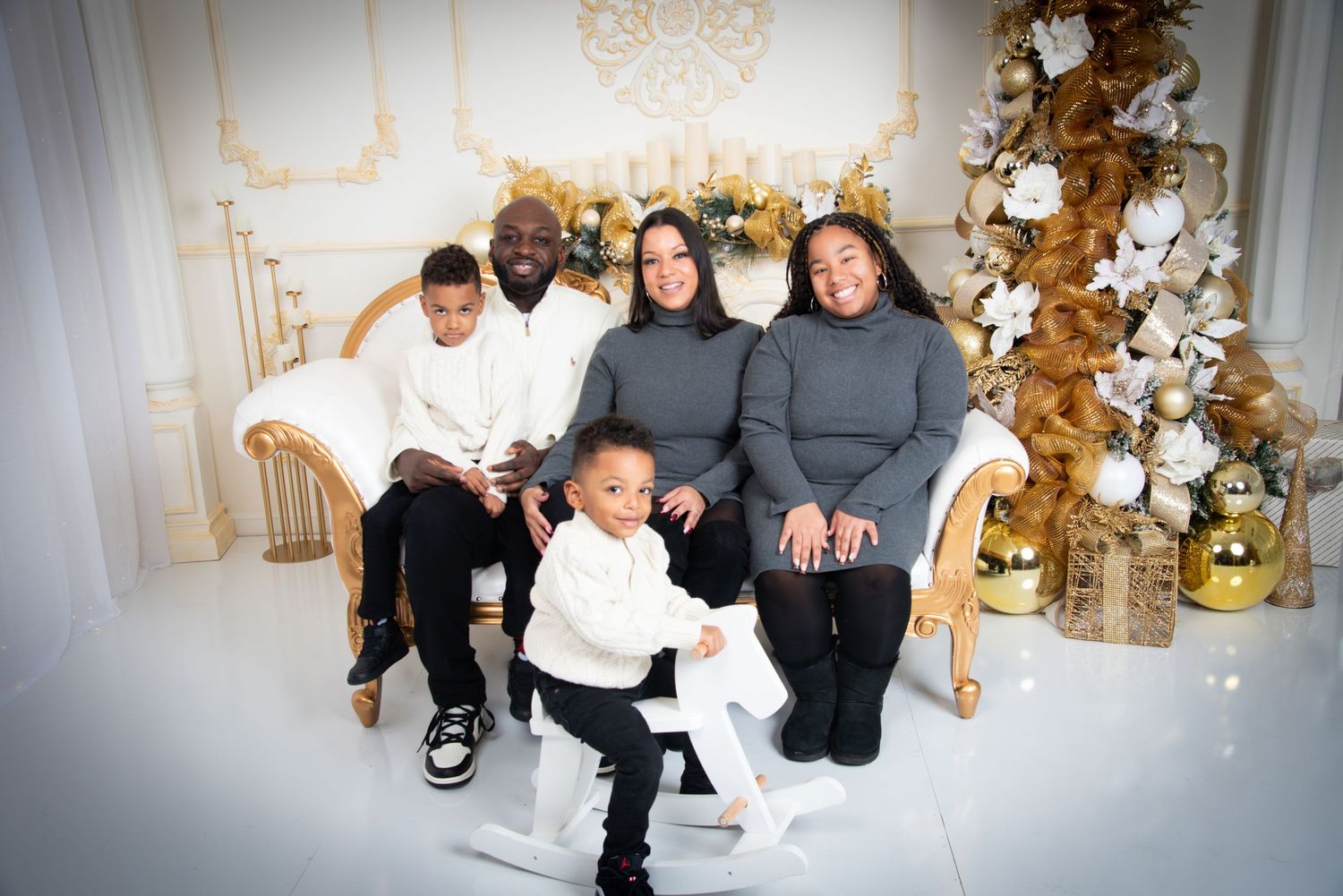 Family of five posing for a festive Christmas portrait in a white and gold holiday decorated studio setting.