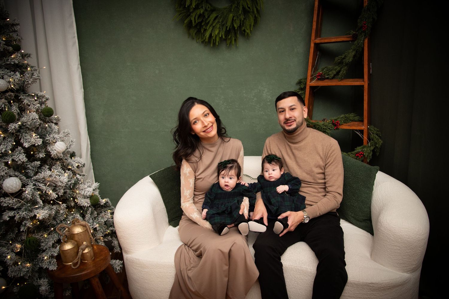Family of four poses on white sofa with Christmas tree and green holiday decor backdrop.