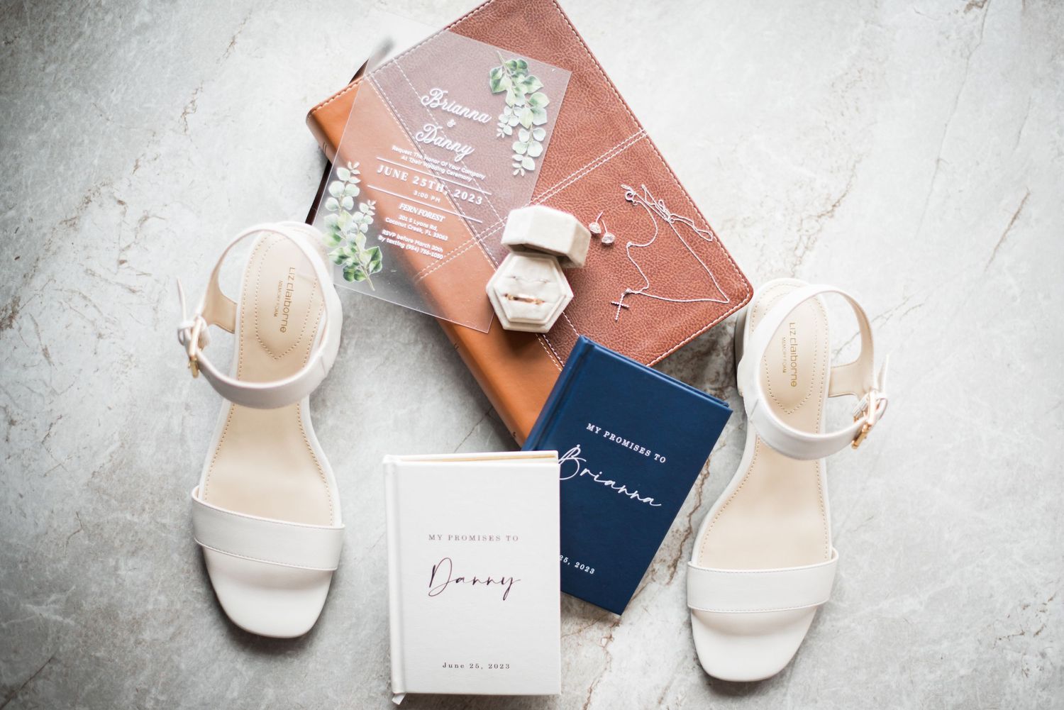 White bridal heels, wedding invitations, vow books, and a ring box styled on a marble surface for a wedding flat lay.