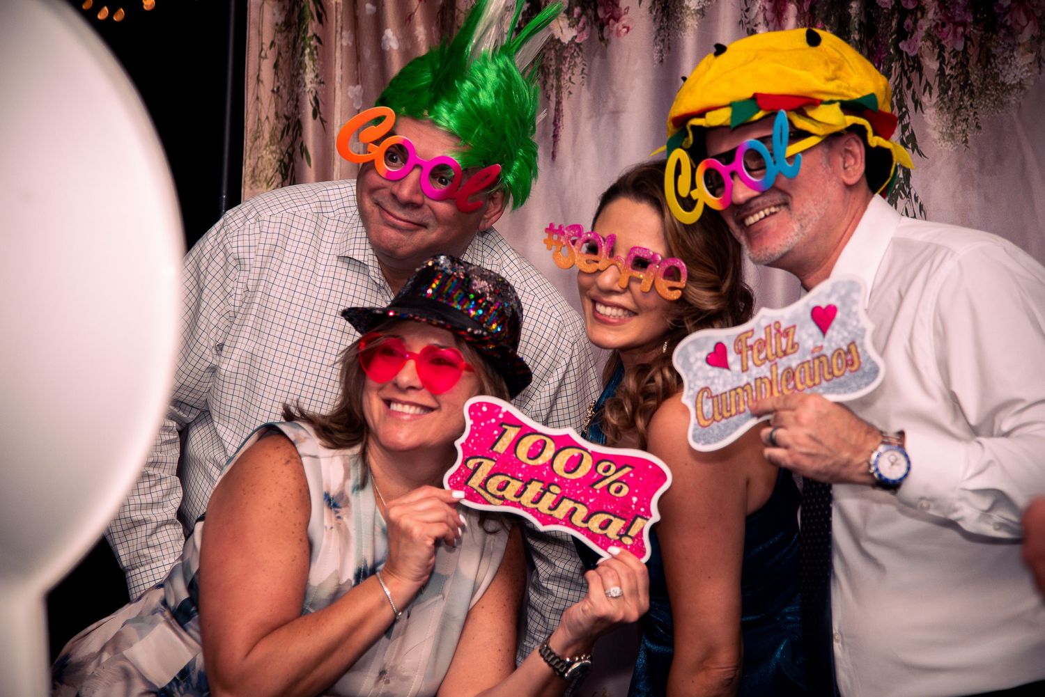 Group of five friends posing in a photo booth wearing fun props, colorful glasses, and hats at a festive party.