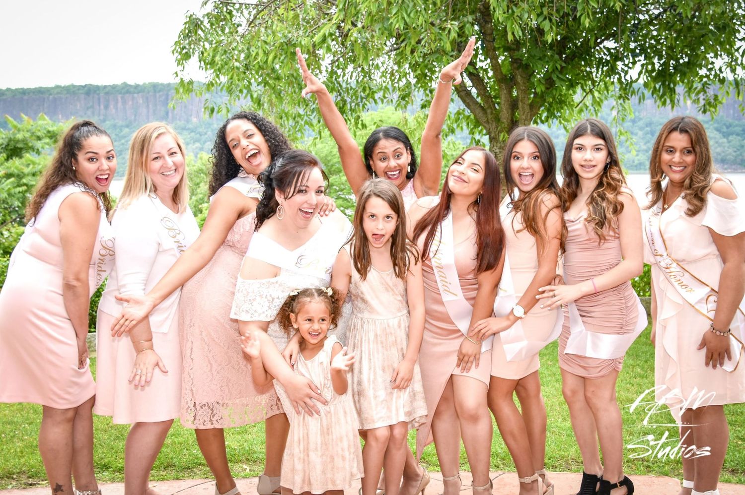 Bridesmaids in matching blush pink dresses pose together outdoors under green trees.