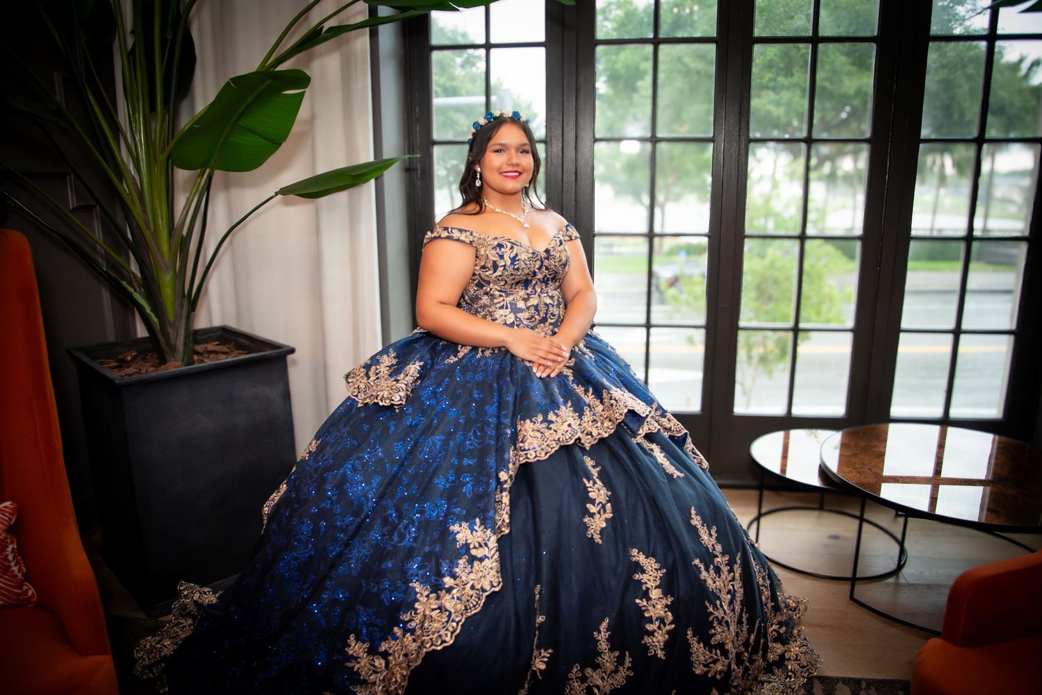 Young woman in navy blue and gold quinceañera ball gown poses elegantly near large windows with garden view.