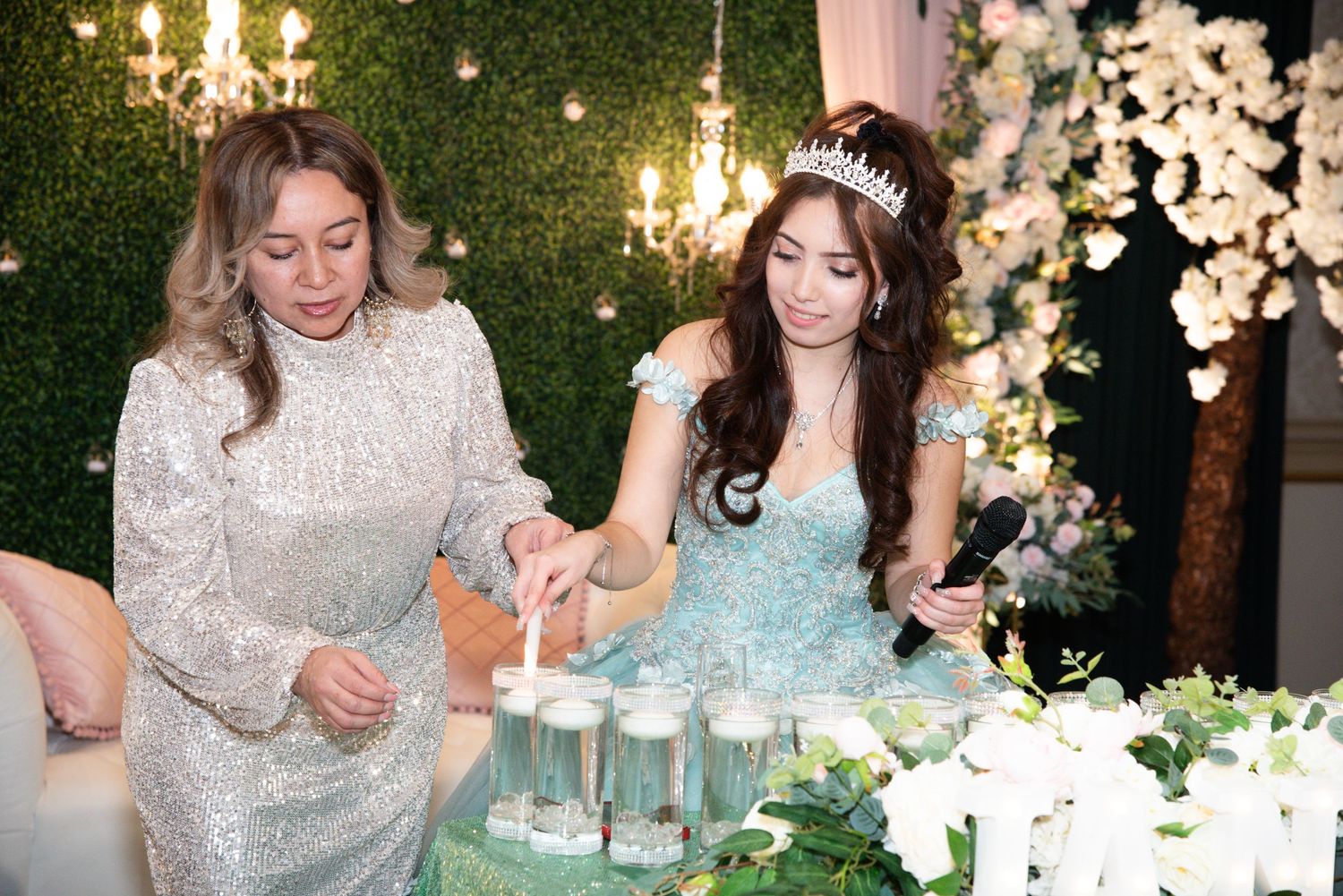 Two people in elegant gowns celebrate at an event with white floral decorations and glowing chandeliers.