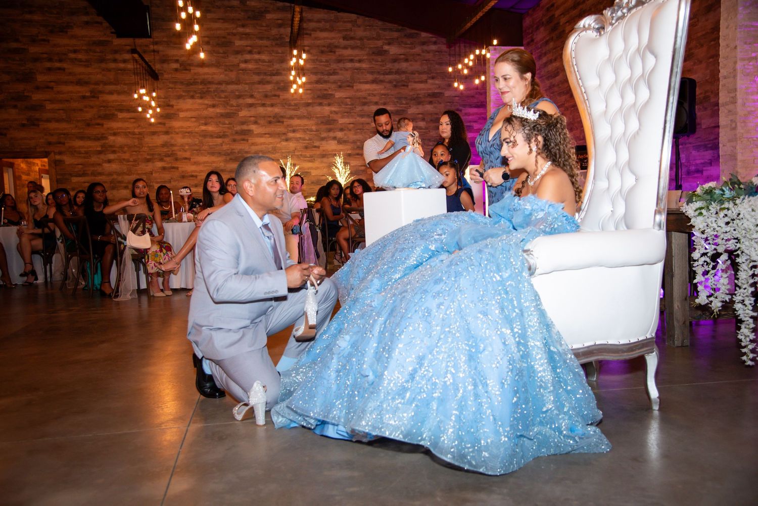 A man kneels to place a shoe on a girl in a blue quinceañera gown seated on a white throne chair at her celebration.