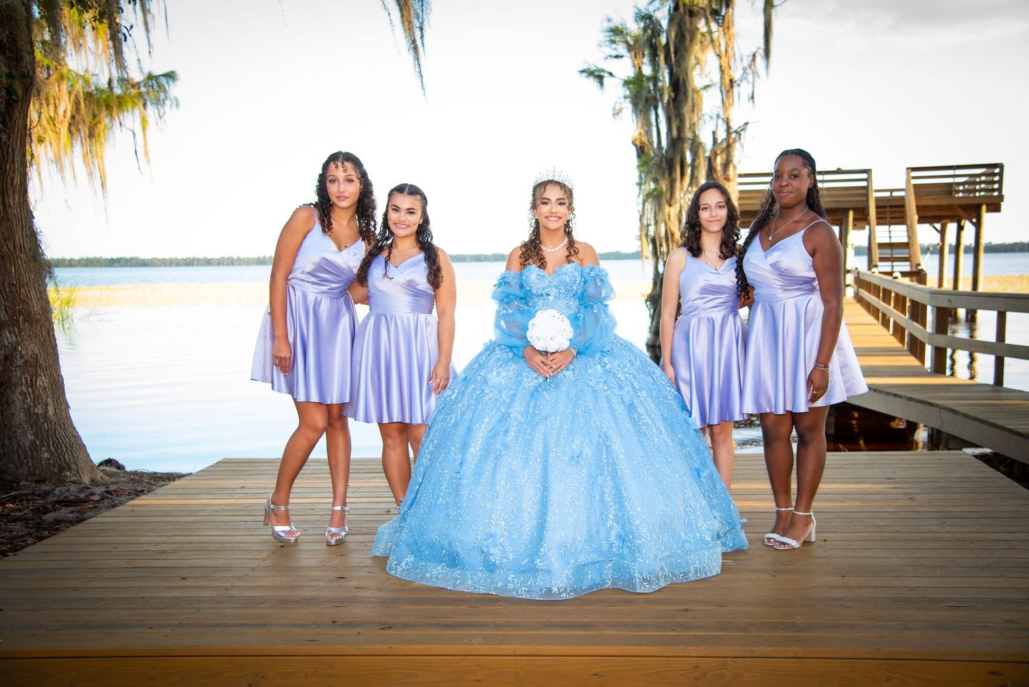 A quinceañera girl in a blue ball gown poses with four bridesmaids in lavender dresses on a lakeside wooden dock.