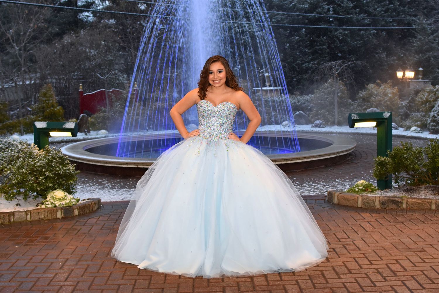 A stunning white ball gown with a sparkly bodice stands in front of an illuminated fountain at dusk.