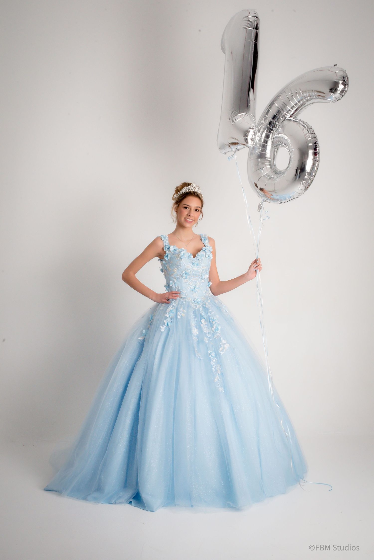Teen girl in light blue ball gown holding silver number 16 balloon, celebrating sweet sixteen birthday.