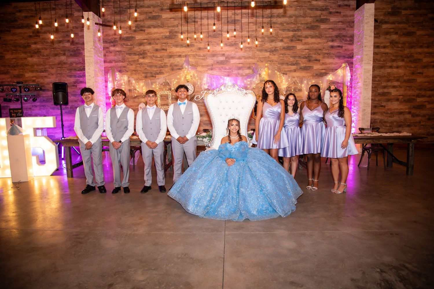 Quinceañera celebration with girl in blue ball gown surrounded by chambelanes and court in elegant decorated venue.