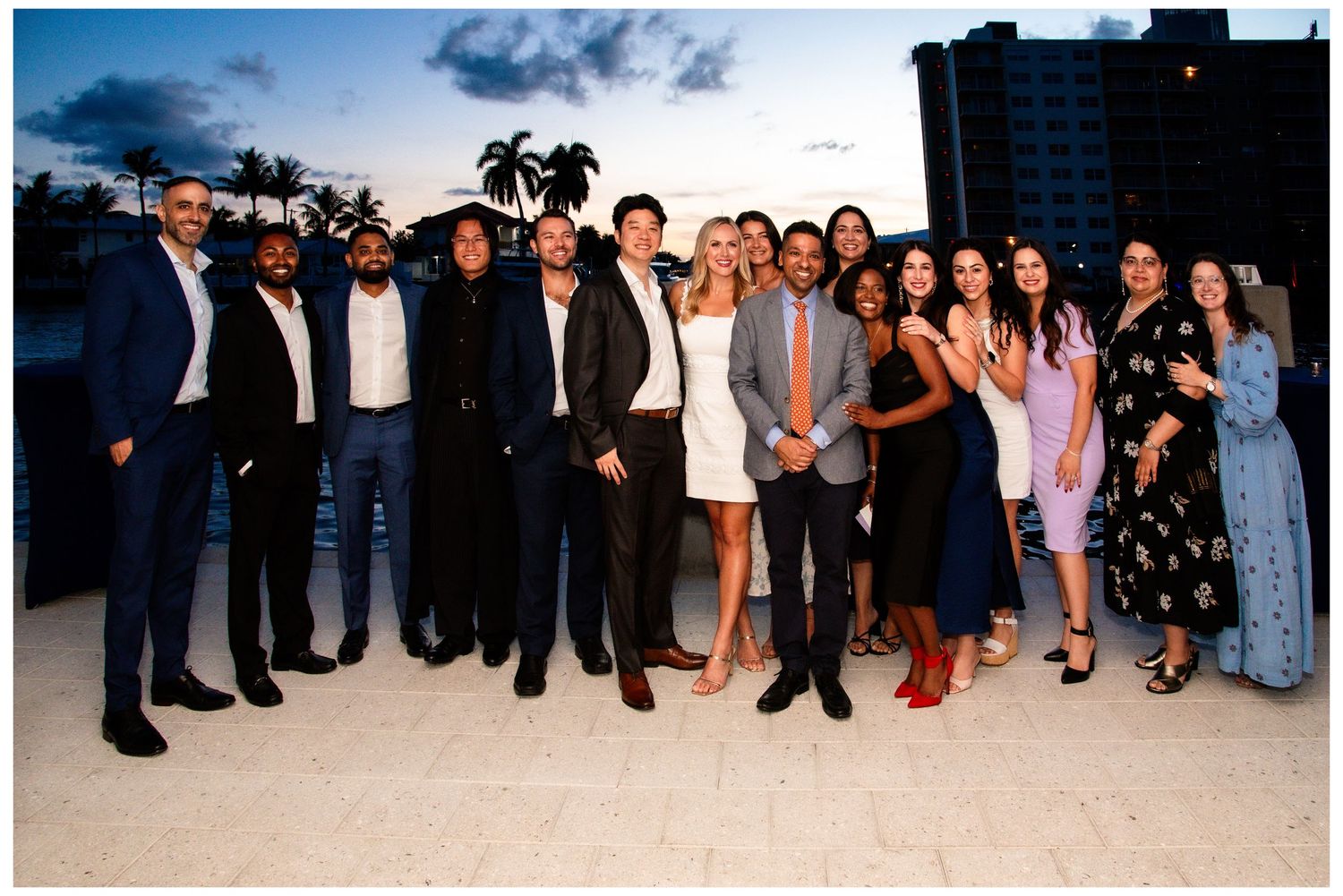 Group of professionals posing together at an outdoor rooftop event during sunset near a waterfront.