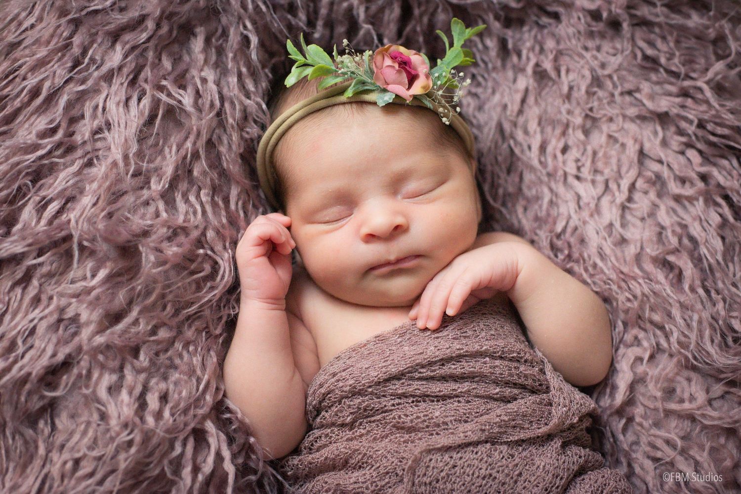 Peaceful newborn baby sleeping on a soft mauve blanket, wearing a delicate floral crown headband.