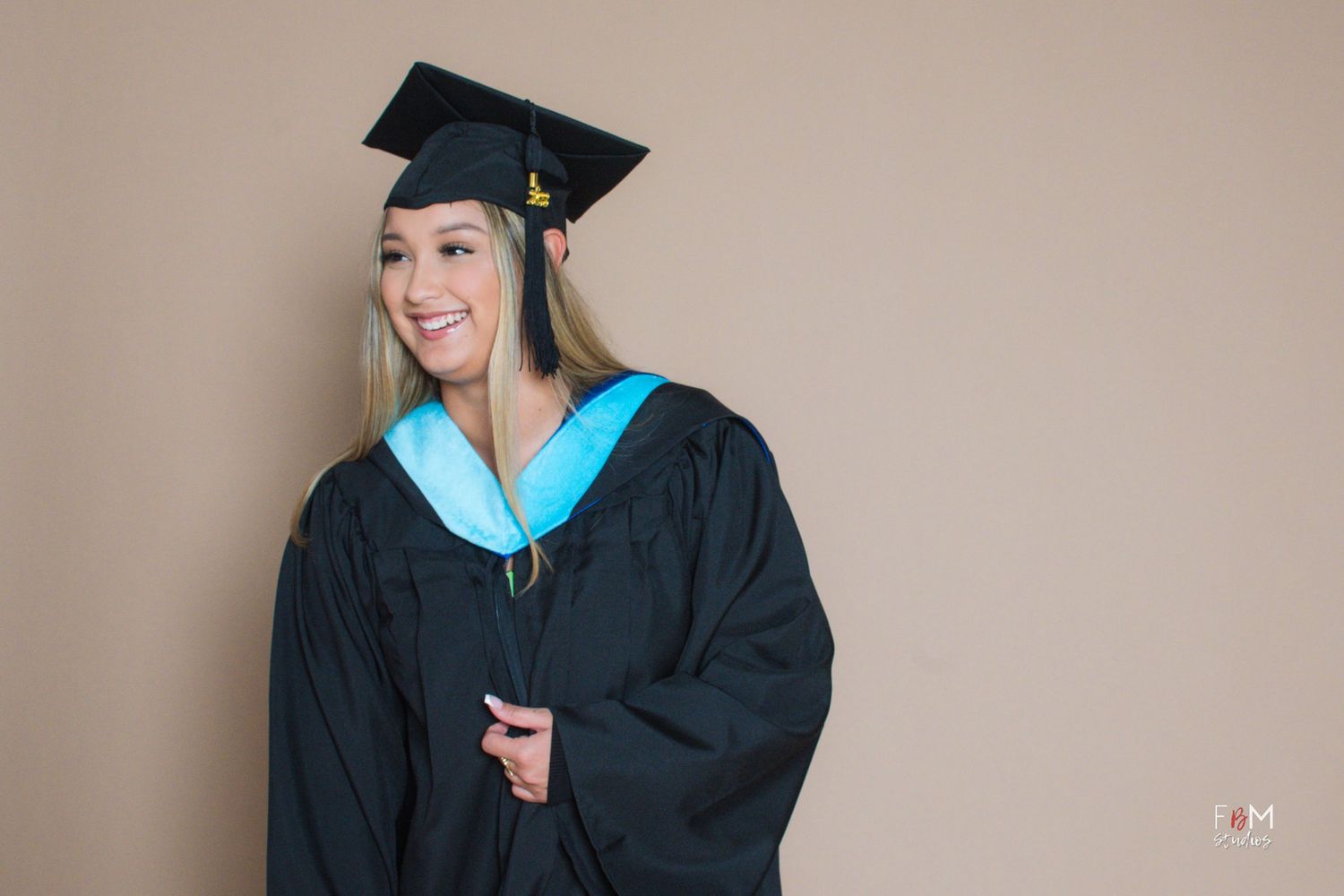 Smiling graduate in black cap and gown with teal hood poses against a beige background.