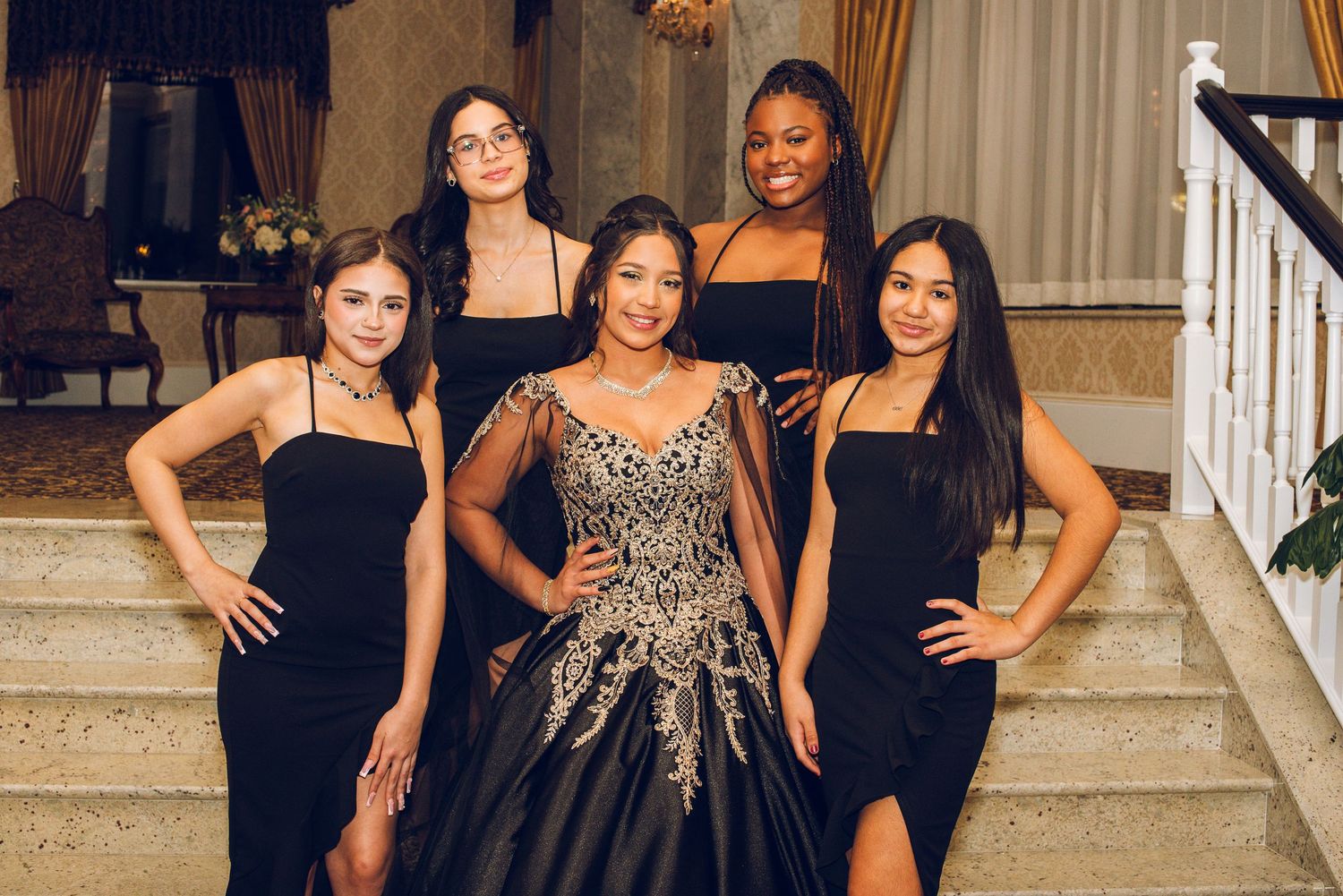 Five young women in elegant black dresses pose on a staircase with one in a gold-embroidered black ball gown centerpiece.