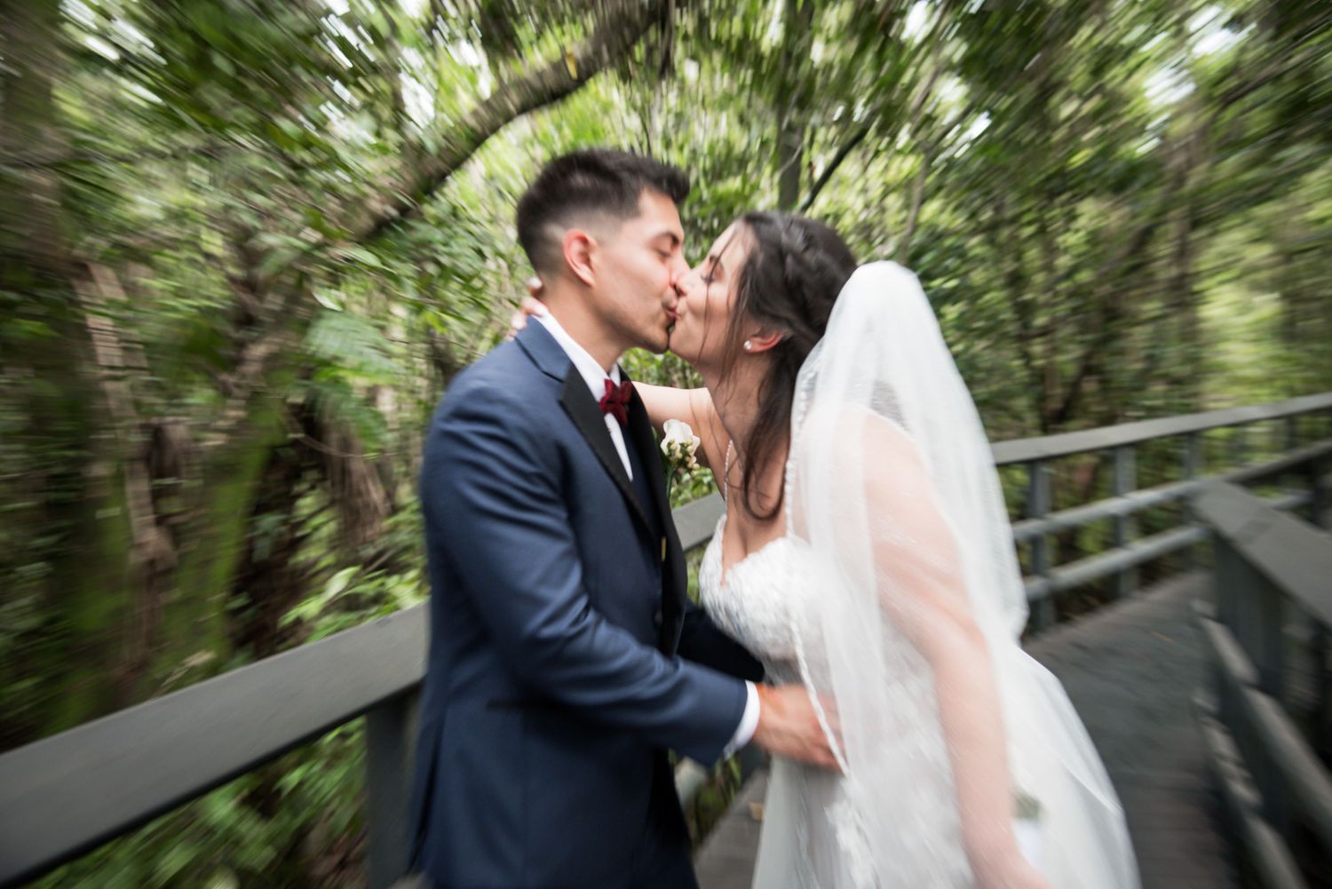 A bride and groom share a romantic kiss on a wooden bridge surrounded by lush green trees on their wedding day.