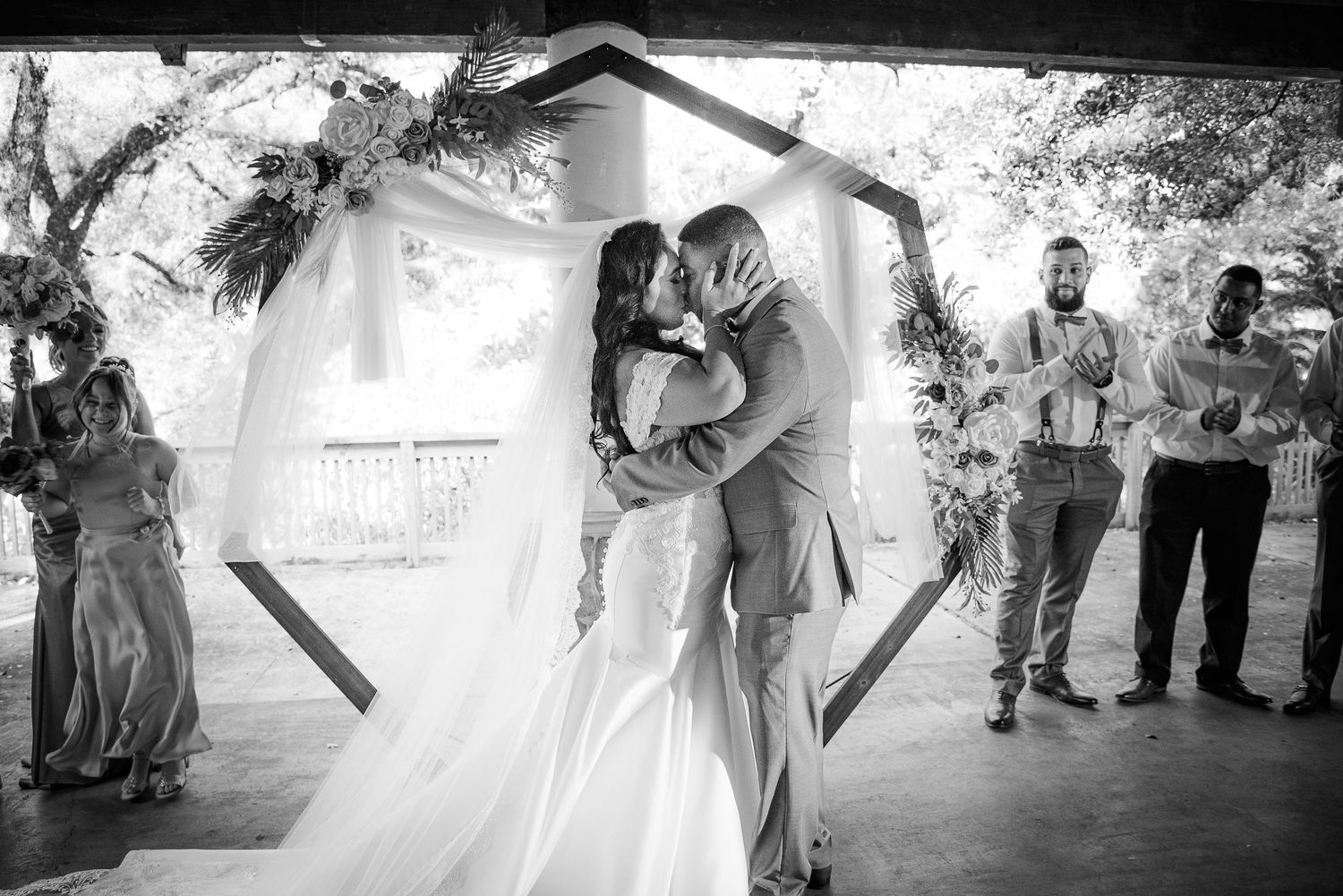 Black and white photo of a bride and groom sharing their first kiss at a wedding ceremony under a hexagonal arch.