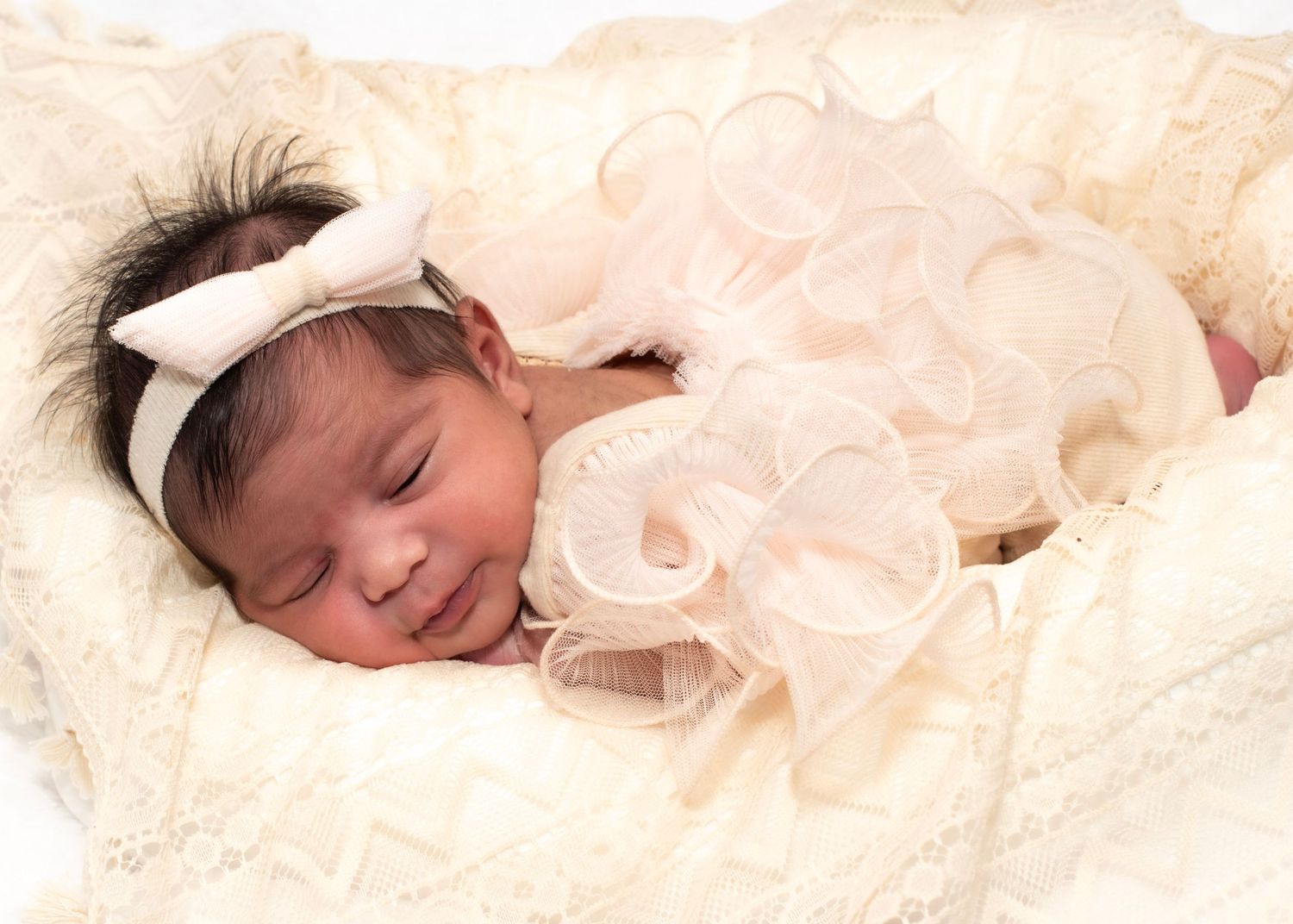 Sleeping newborn baby girl in cream lace outfit with bow headband, peacefully resting on soft white blanket.
