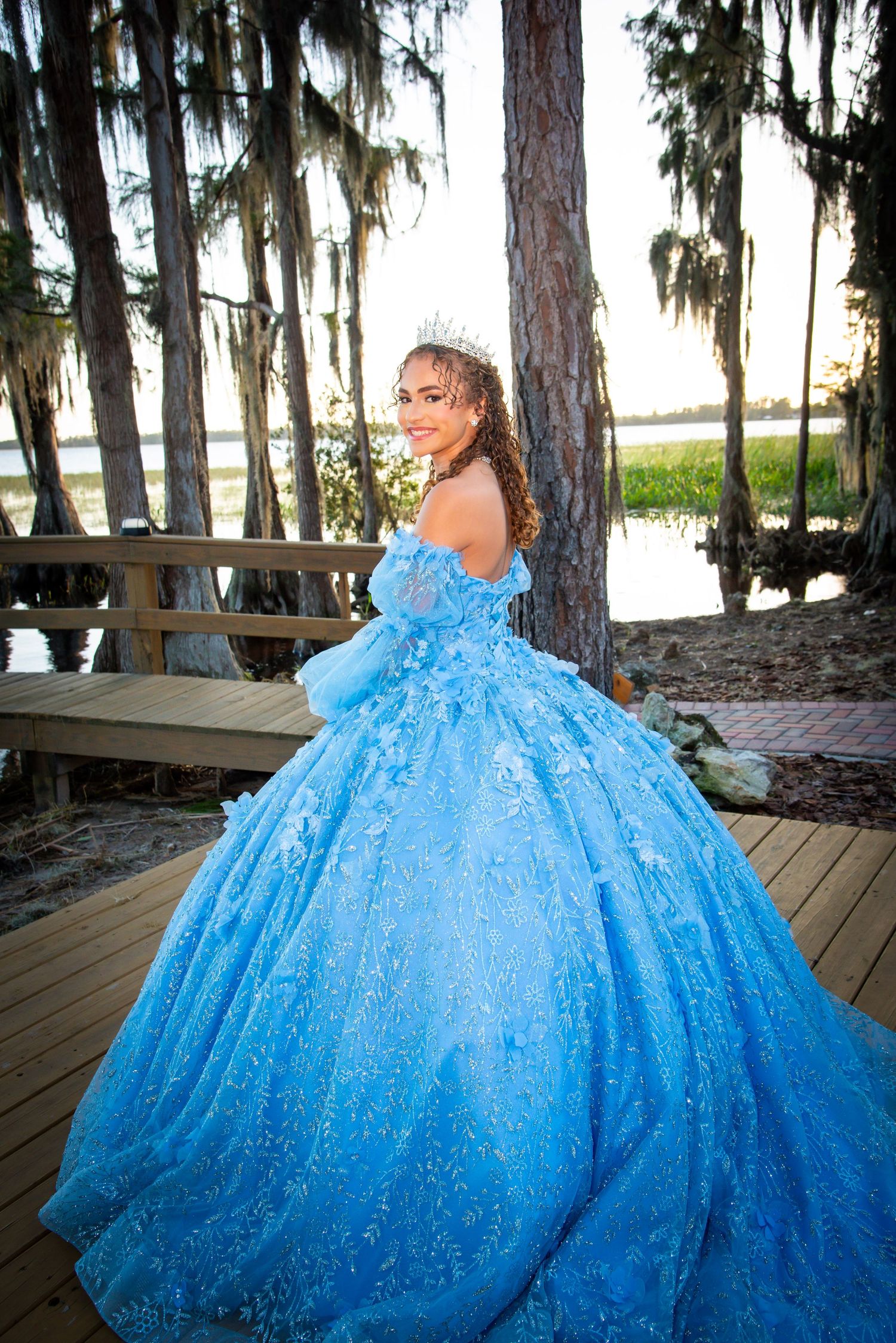 Young woman in a stunning blue ball gown with floral details and tiara poses on a wooden deck.