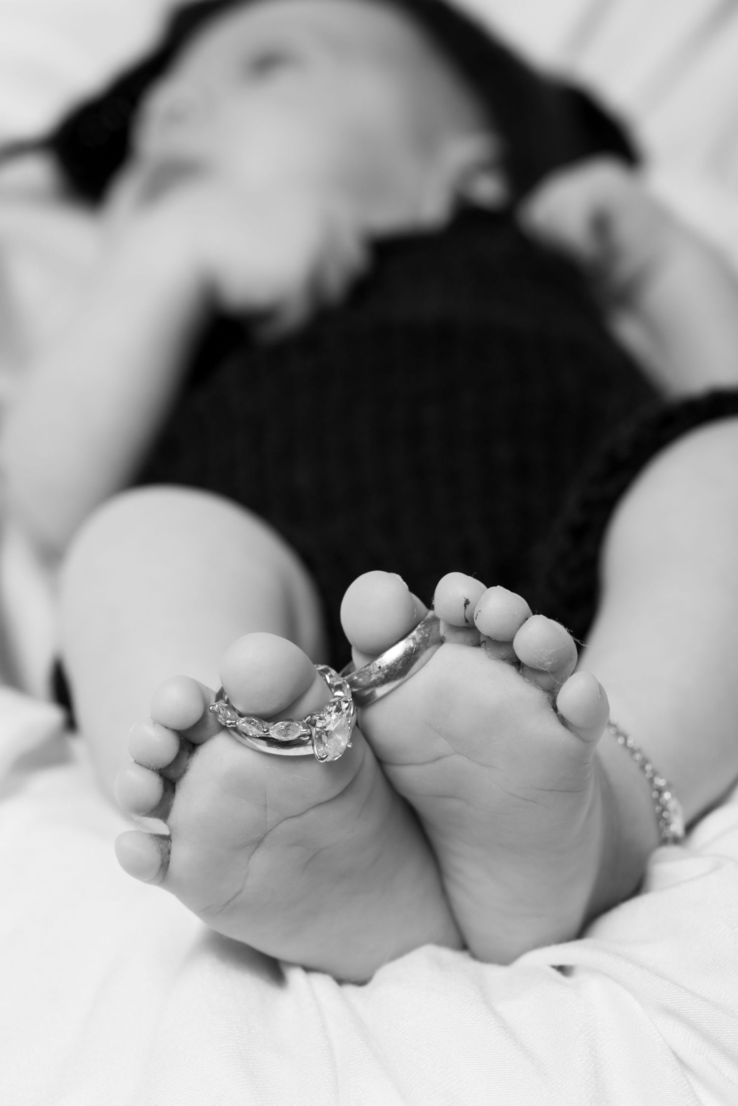 Close-up black and white photo of sleeping baby feet adorned with delicate jewelry rings and bracelets.