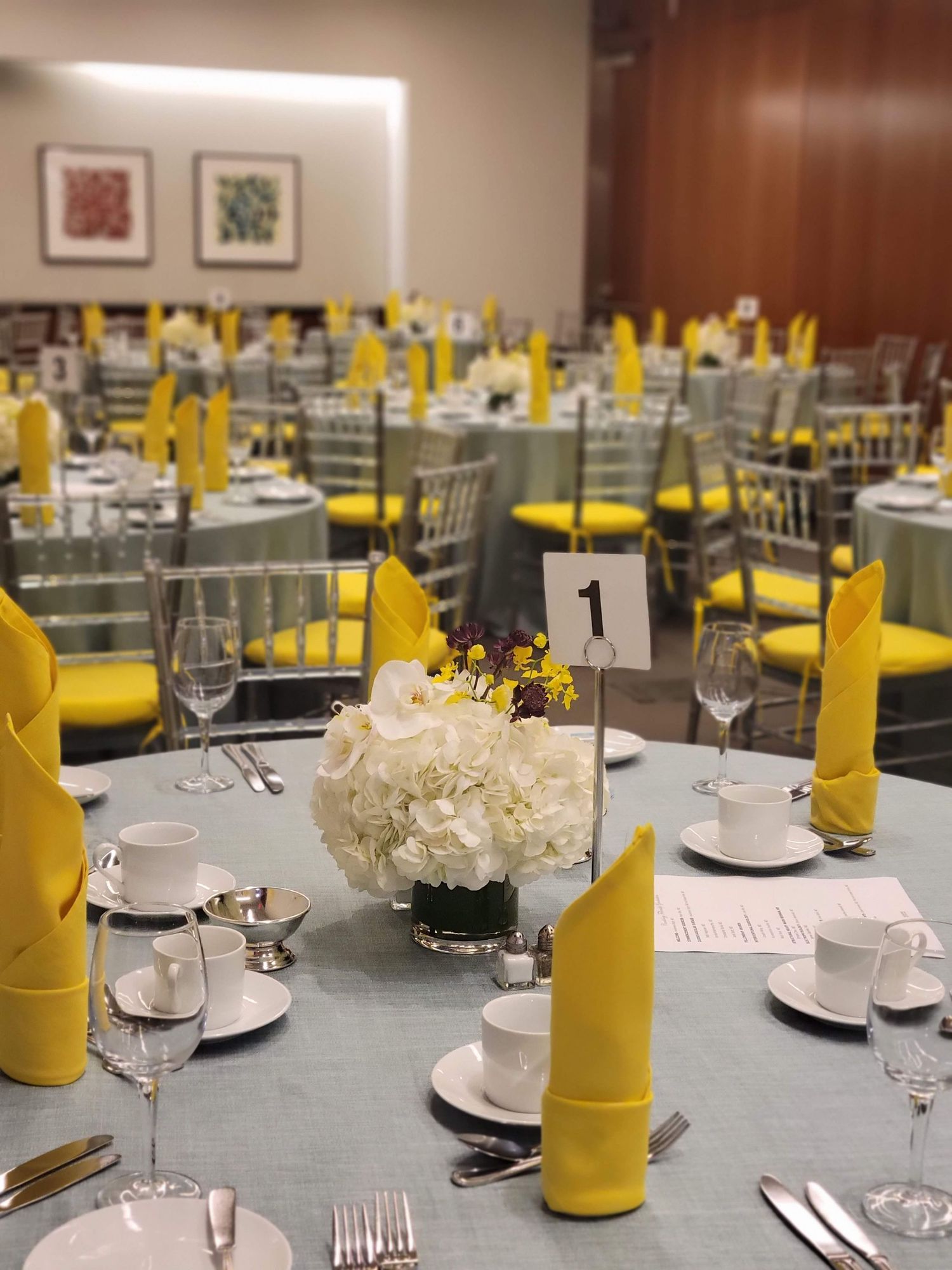 Elegant banquet hall with round tables set with yellow napkins, white hydrangea centerpieces, and gray linens.
