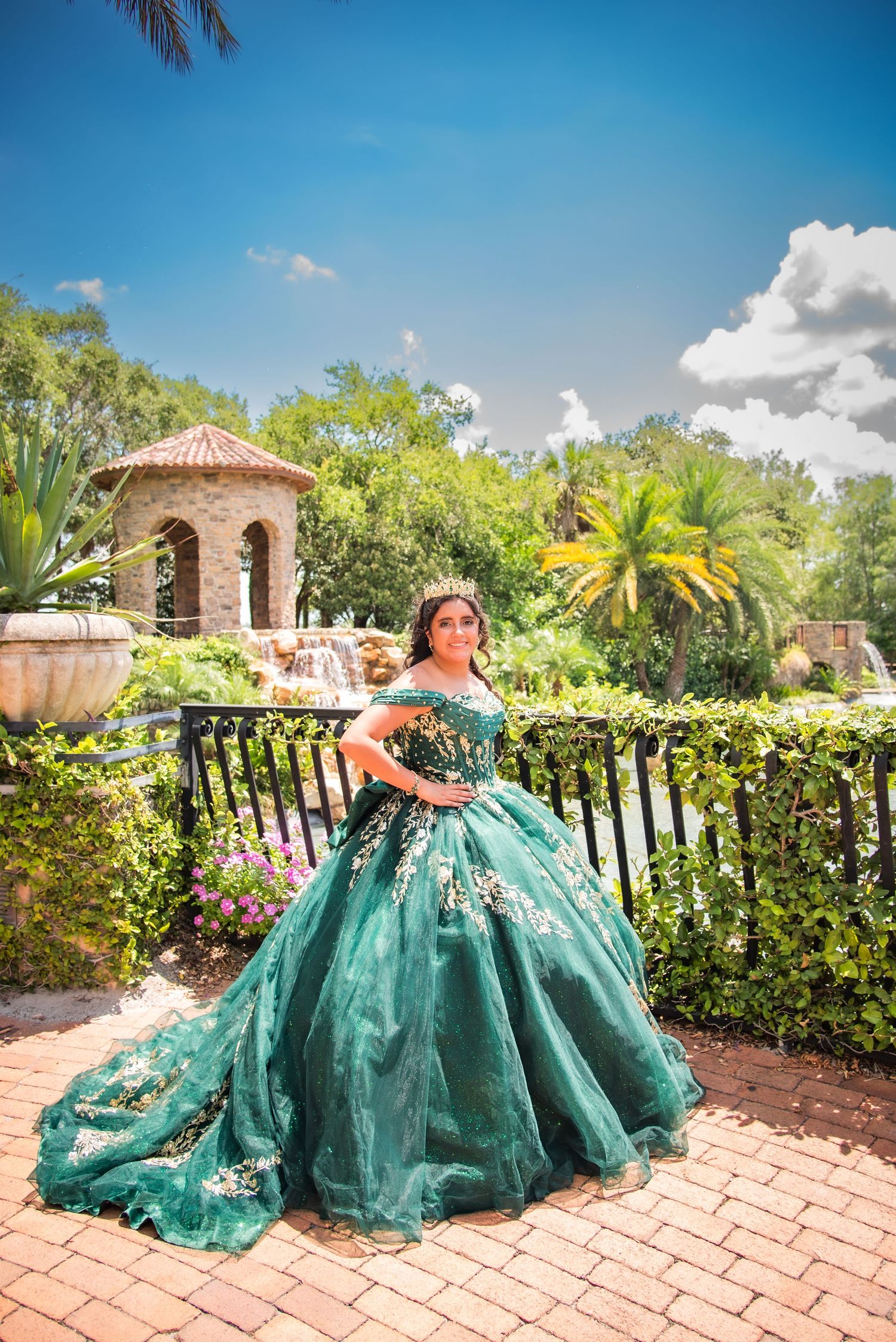 Young woman in elegant emerald green ball gown with floral details poses in a lush garden for quinceañera.