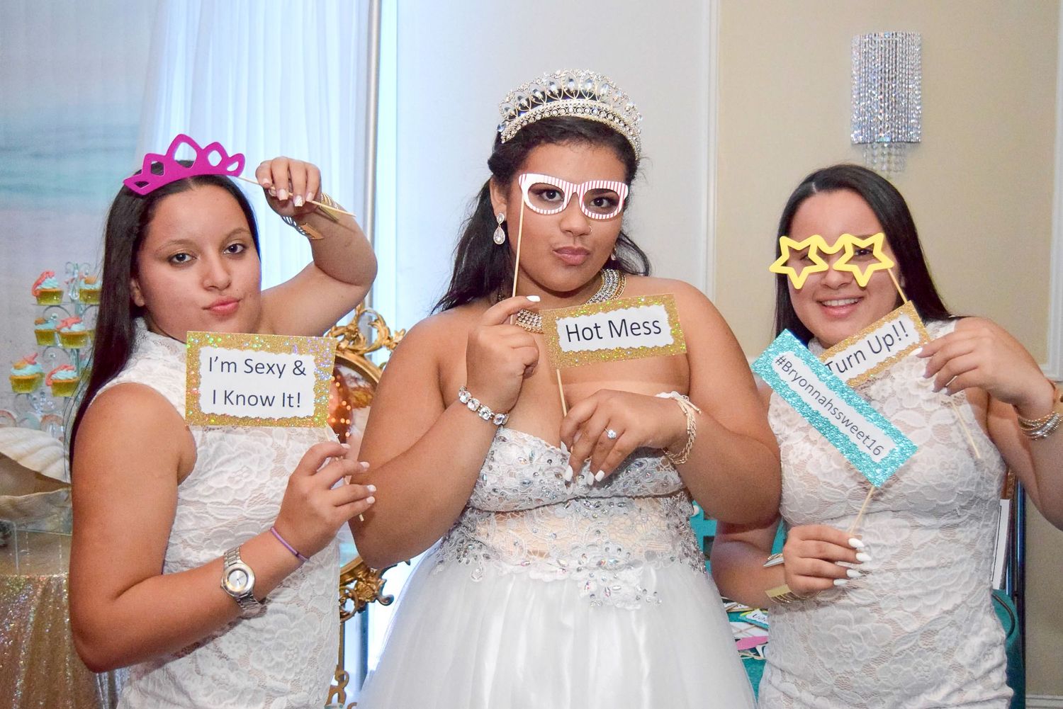 Three women pose with fun photo booth props at quinceañera celebration, holding signs and novelty glasses by FBM Studios in Fort Lauderdale