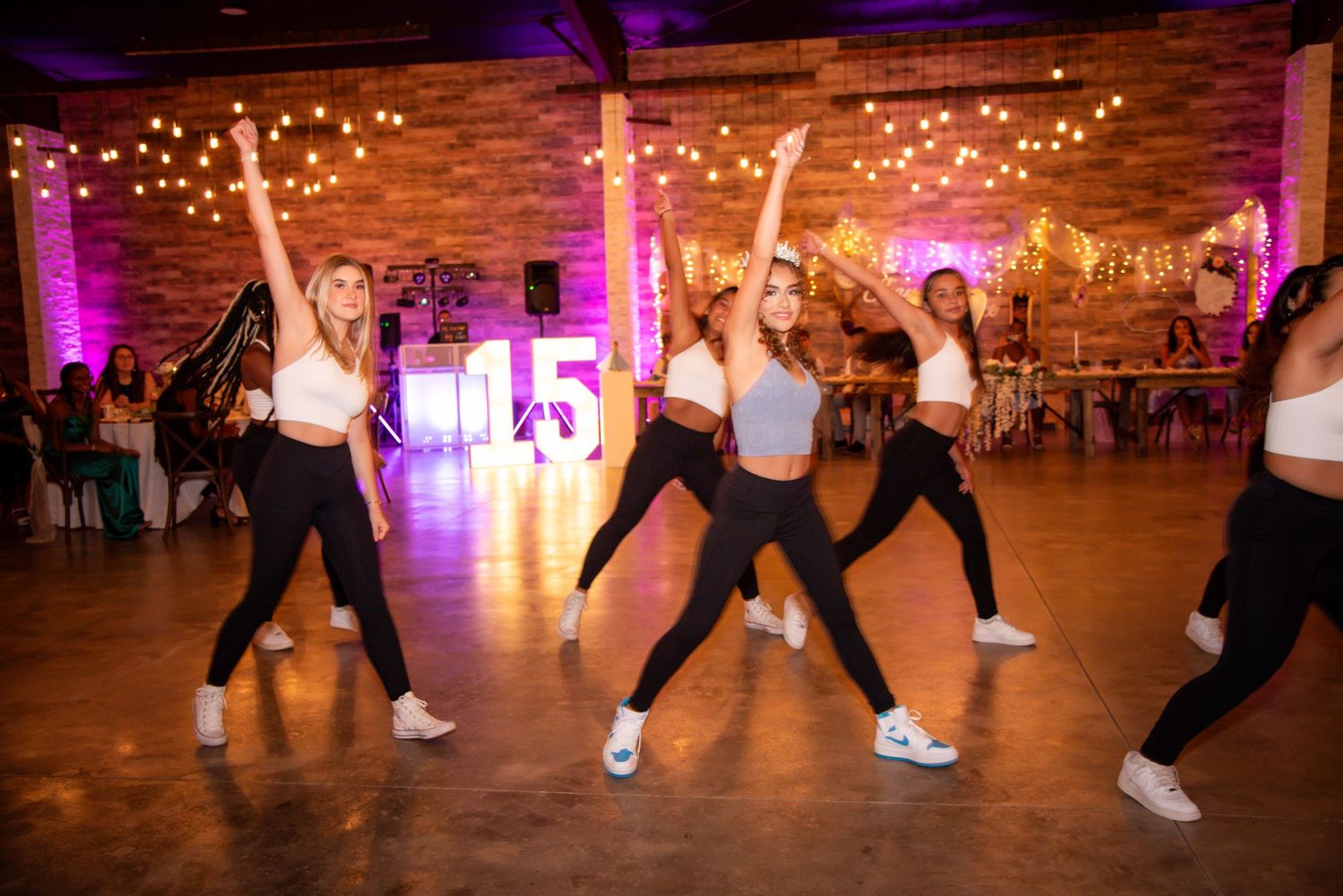 Young women dancing in white crop tops and black pants at a festive quinceañera celebration with purple lighting.