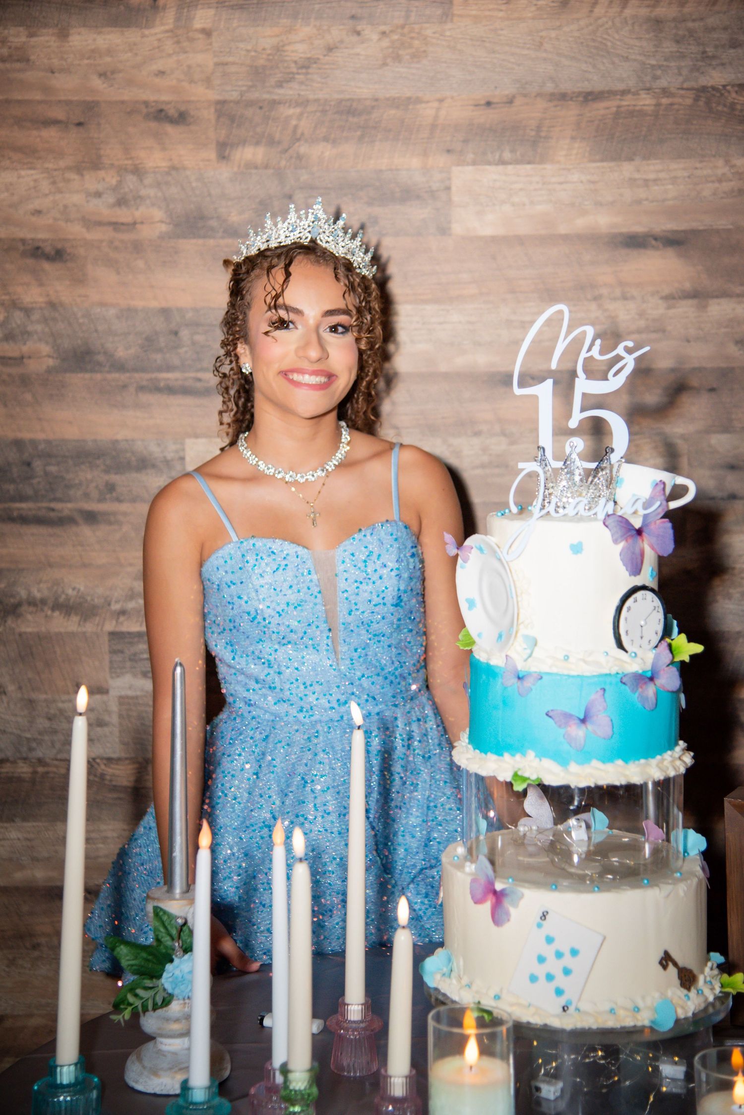 A young woman in a blue sequin dress and tiara poses beside her quinceañera cake celebrating her 15th birthday.