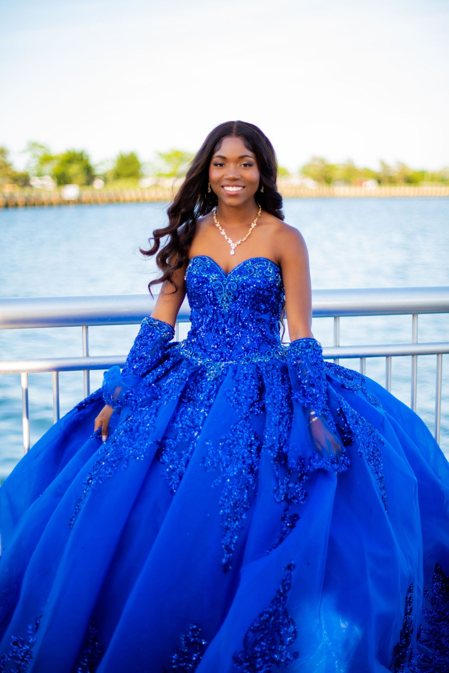 Young woman in royal blue sequined ball gown with gloves poses by waterfront railing, smiling elegantly.