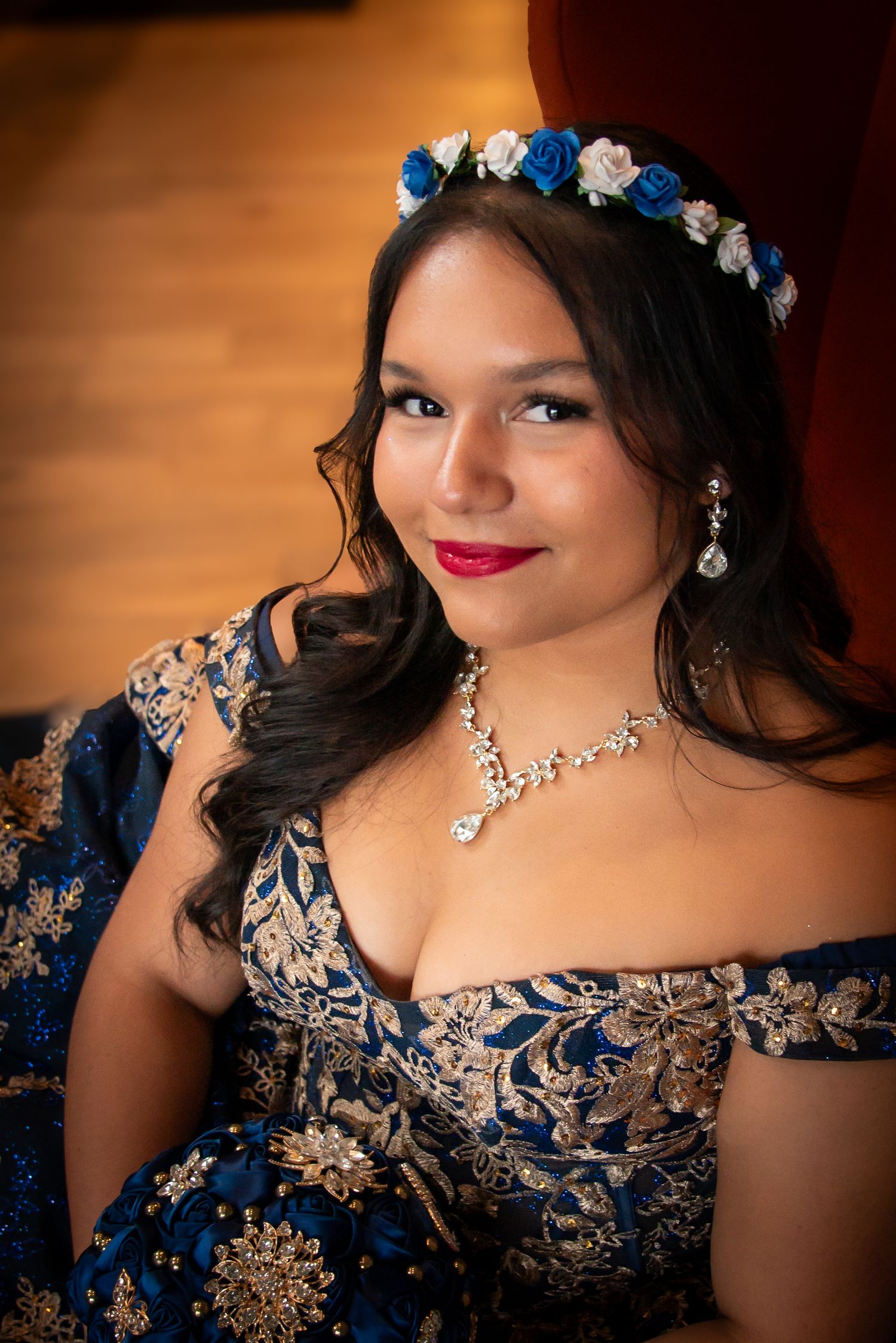Young woman in blue floral off-shoulder gown with floral crown, diamond necklace, and red lips poses elegantly.
