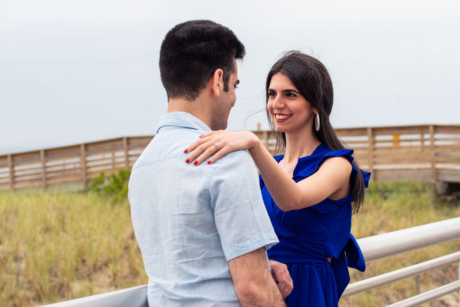 A smiling woman in a blue dress places her hand on a man's shoulder outdoors near a wooden boardwalk.
