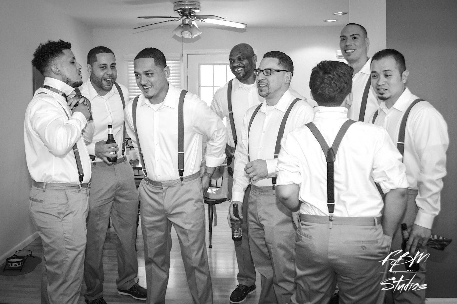 A black and white panoramic photo showing groomsmen in suspenders and dress shirts laughing together at a wedding.