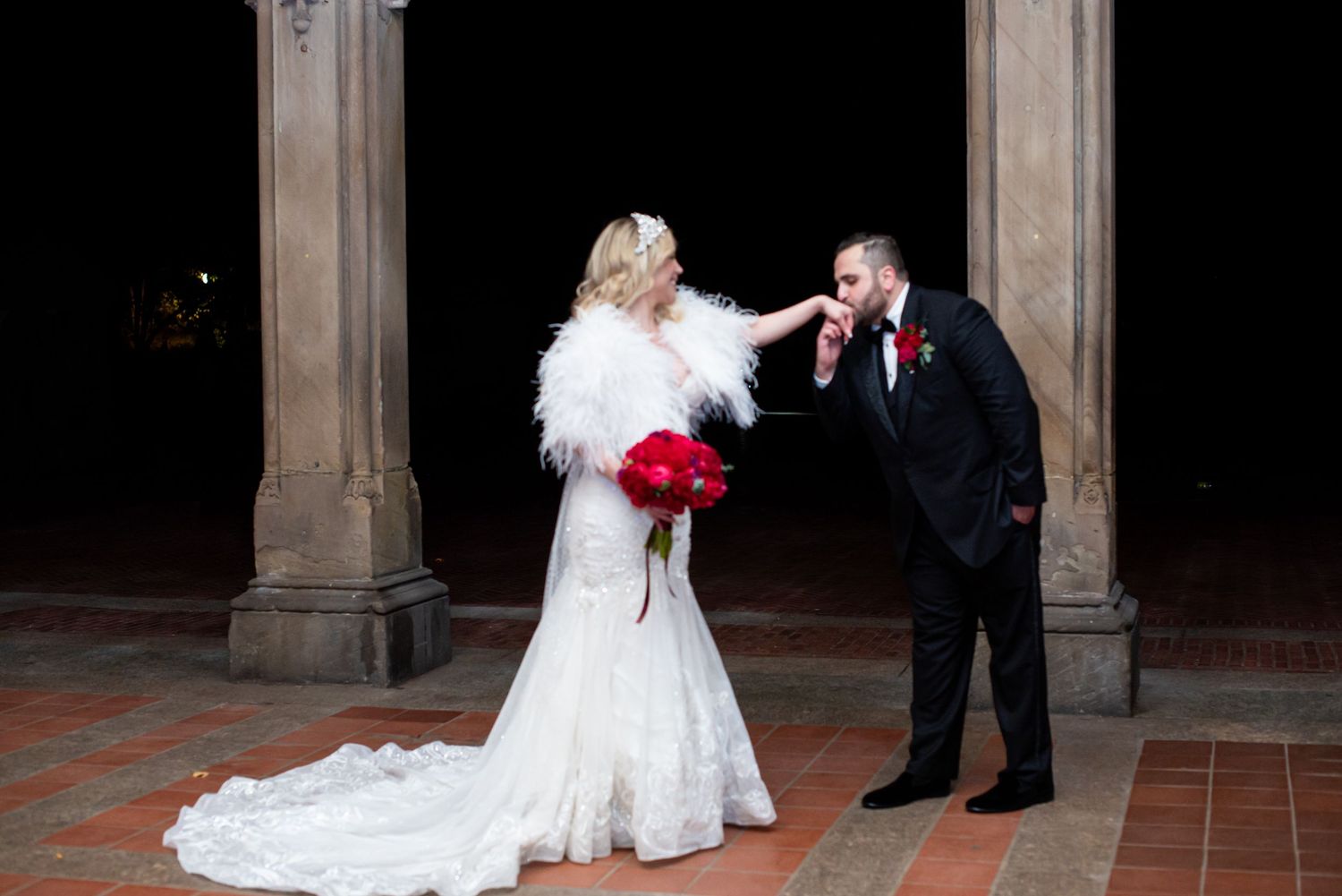 Groom in black tuxedo kissing bride's hand outdoors at night, bride wearing white lace gown and feather wrap holding red bouquet.