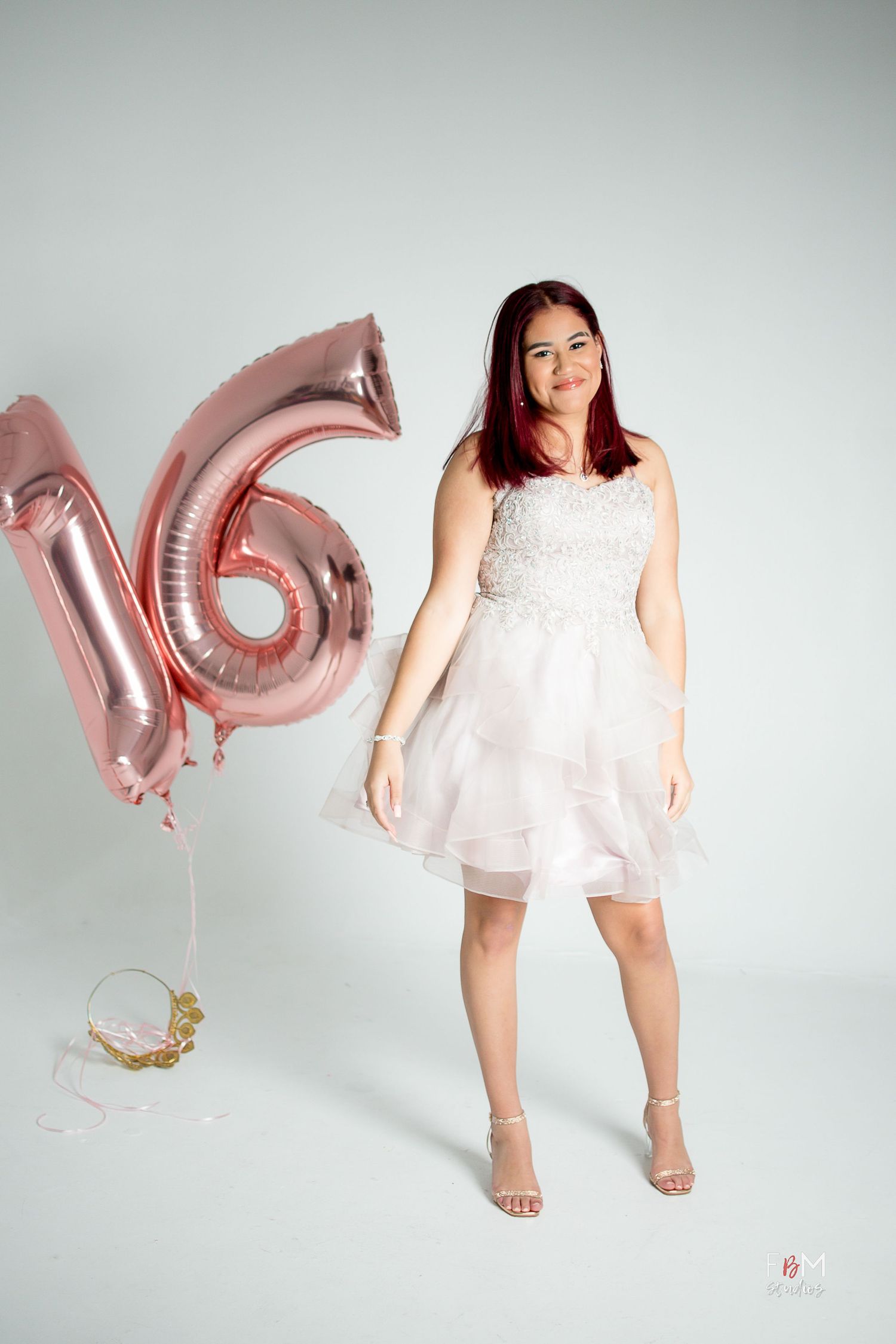 A teenage girl in a white dress poses next to rose gold number 16 balloons for her sweet sixteen celebration.