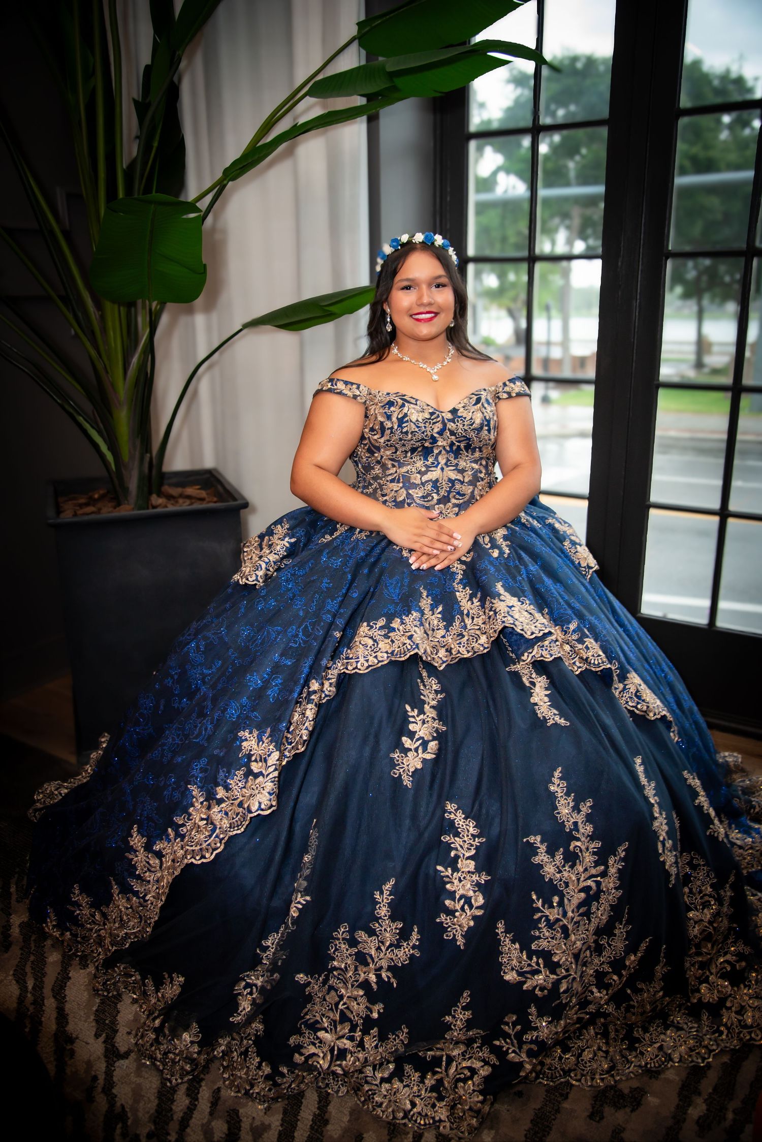 Young woman in navy blue and gold quinceañera ball gown with floral crown poses elegantly indoors.