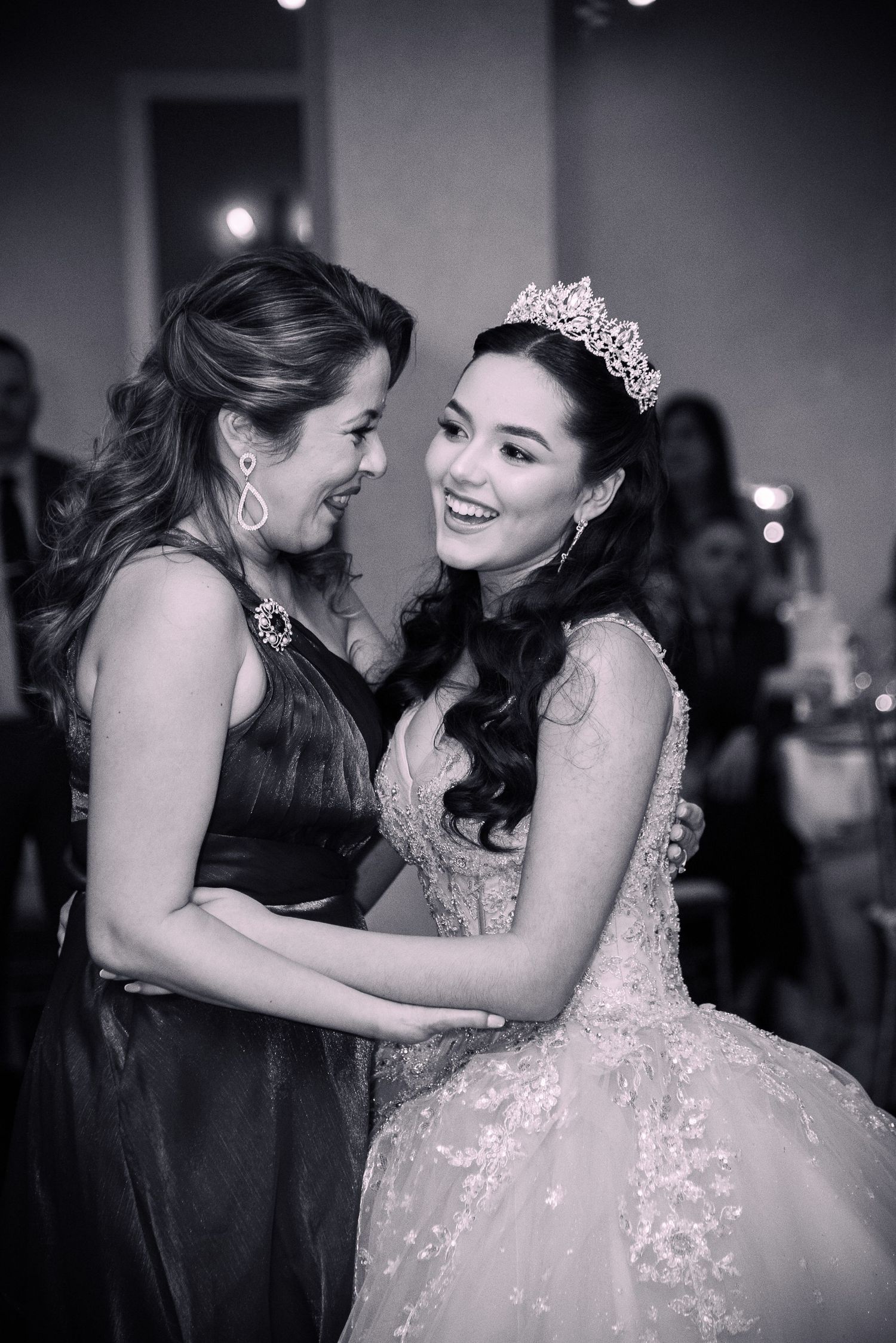 Black and white photo of two women smiling and embracing at a quinceañera celebration, one wearing a tiara and ballgown by FBM Studios in FL