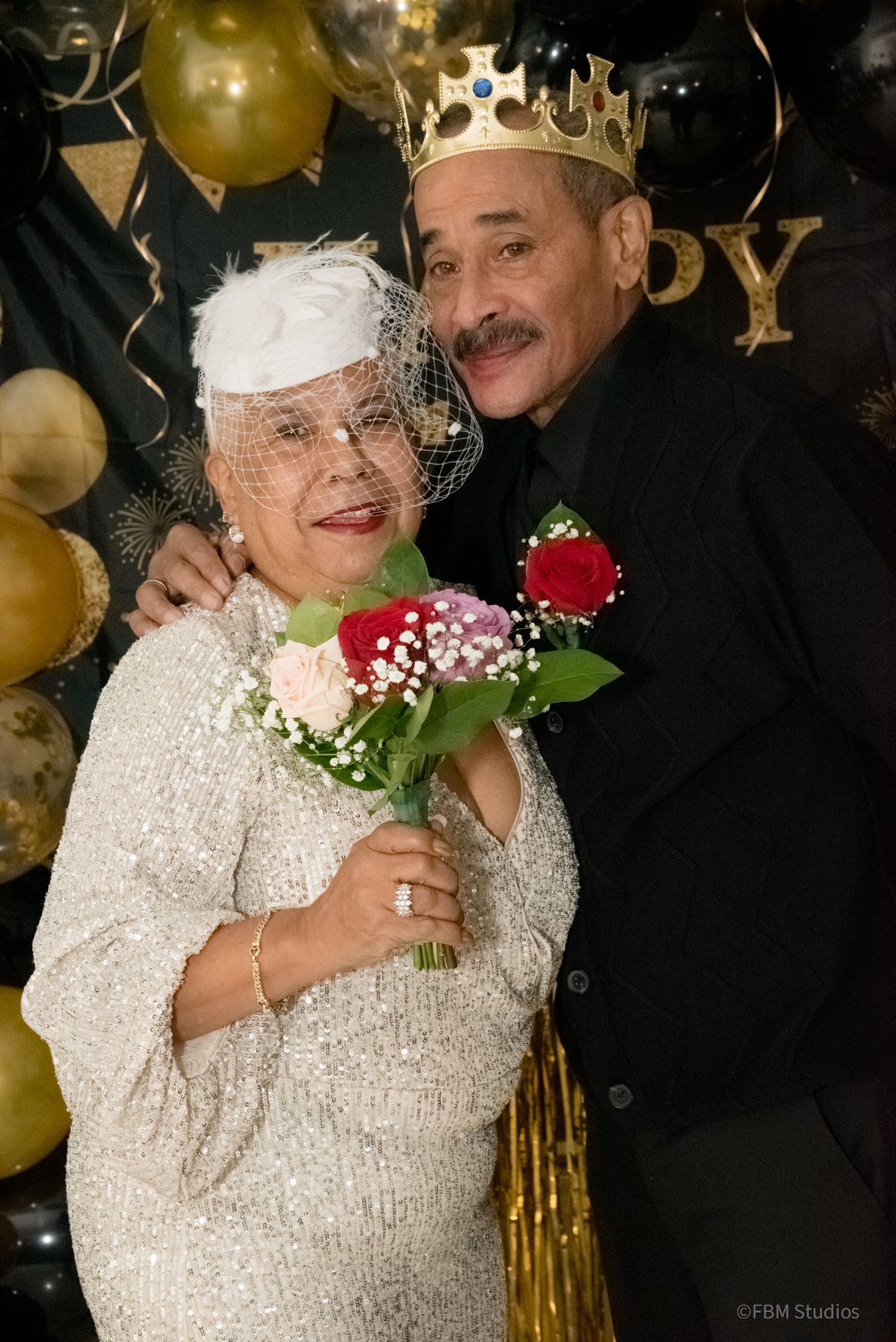 A couple celebrates at a party with gold balloons and confetti while wearing formal attire and holding flowers by FBM Studios in Fort Lauder
