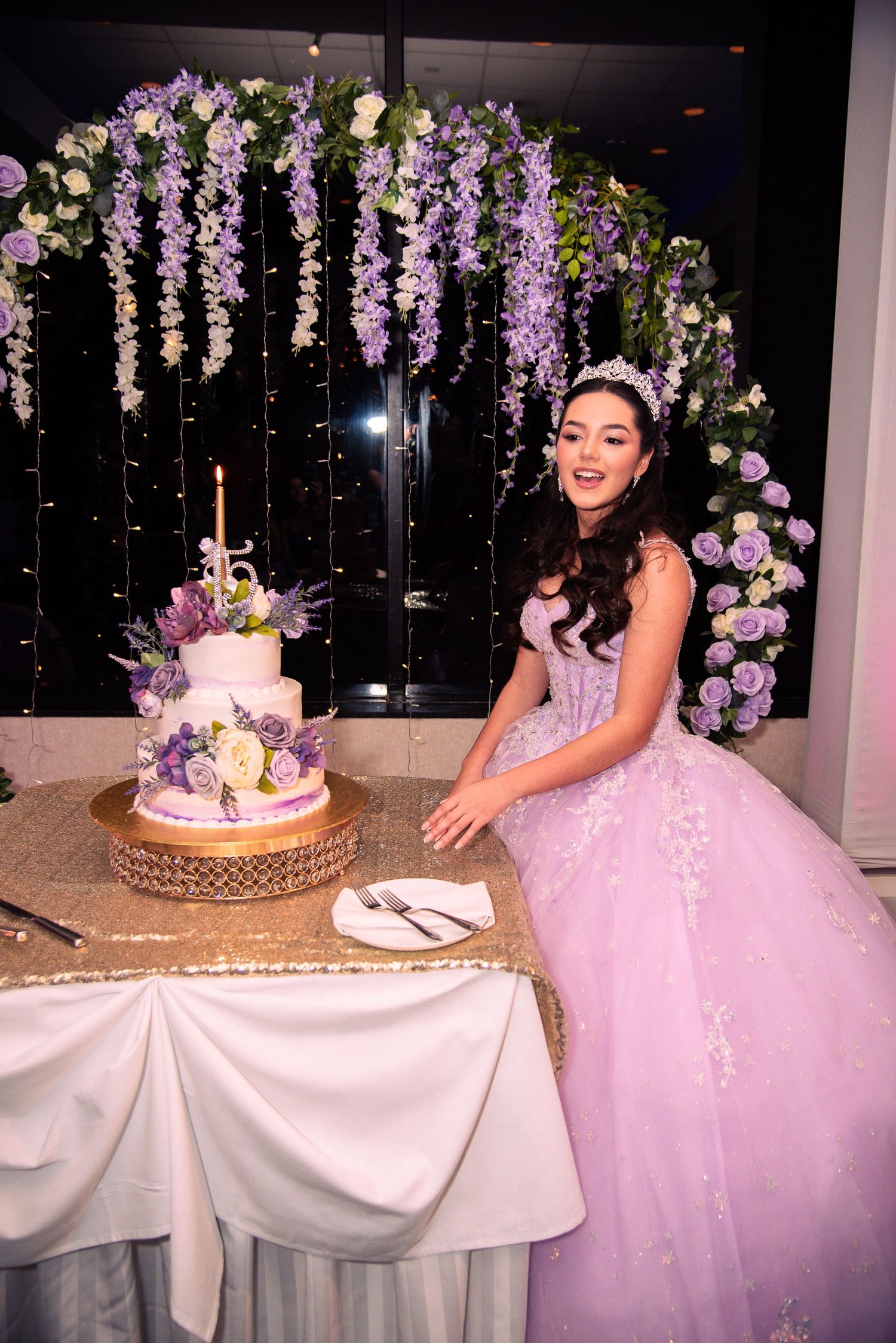 Young woman in lavender quinceañera gown poses beside tiered birthday cake under floral arch with purple flowers.