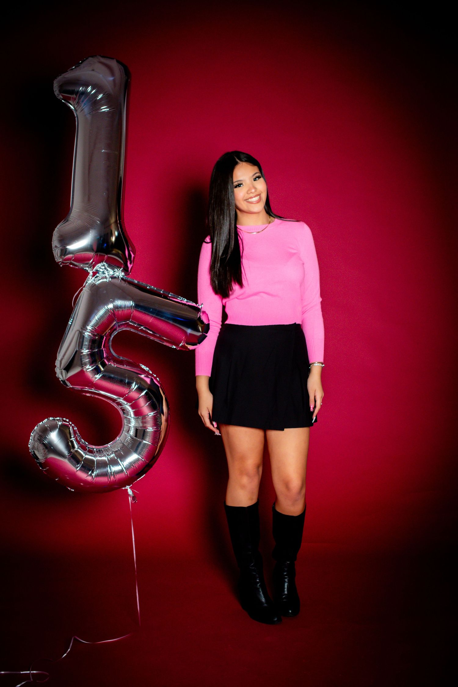 Young woman in pink top and black skirt poses beside silver number 15 balloons against red background.